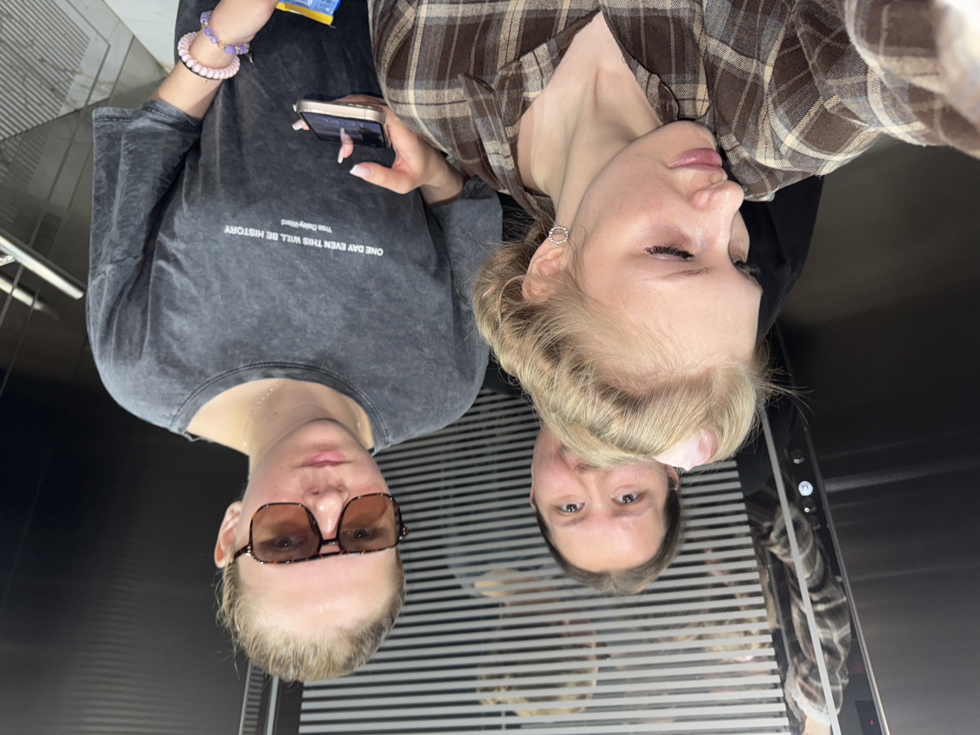Three young women are taking a selfie in what appears to be an elevator. The woman in the foreground, on the left, is looking down and to the left, wearing a brown and tan plaid shirt. The woman in the middle is smiling and looking at the camera, her head is slightly tilted. The woman on the right is wearing large, brown-tinted sunglasses and a dark grey t-shirt with white text that reads "ONE DAY EVEN THIS WILL BE HISTORY Yrsa Daley-Ward". She is holding a smartphone in her right hand and wearing a pink and purple bracelet on her left wrist. The elevator walls are metallic, with a mirrored surface reflecting the subjects and the elevator interior, including a set of horizontal blinds. A handrail is visible in the background to the right. There is no indication of the specific location within the city of Perm, Russia.