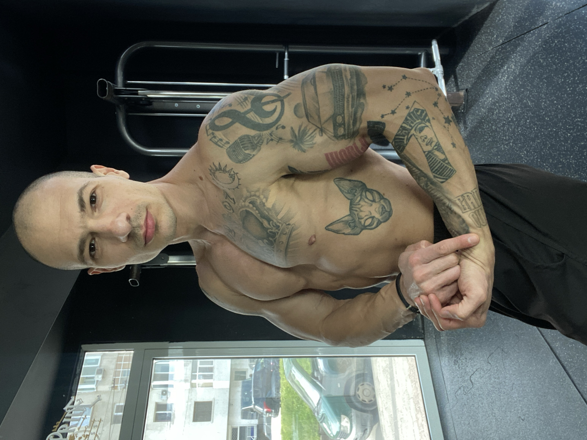 A muscular man with a shaved head and extensive tattoos is posing indoors, likely at a gym, given the equipment in the background. He is shirtless, flexing his arms, and wearing black shorts or pants. The environment includes a window with a view of a building and cars, possibly placing the location in Pleven, Bulgaria. The floor is a speckled grey. The walls are black.