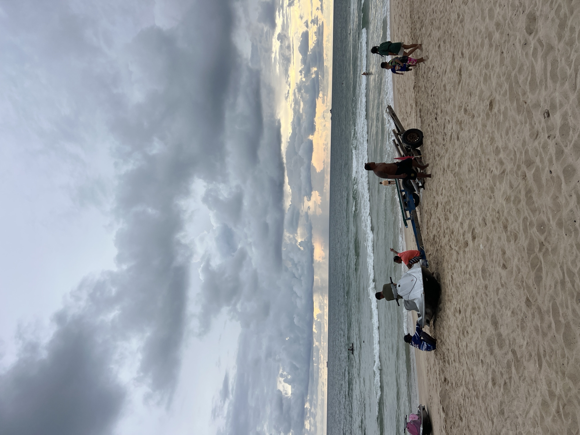 A group of people are on a sandy beach near the ocean. The sky is filled with dramatic, dark clouds, suggesting an overcast or stormy weather condition. The sun is partially visible through a break in the clouds, casting a soft, warm light that creates a contrast with the darker areas. The waves are gently rolling onto the shore, indicating a calm sea despite the cloudy sky.
Several people are scattered across the sand. Some are walking along the beach, while others are gathered around a jet ski that is being transported on a trailer. The jet ski is white and black. One person is standing near the trailer, appearing to be supervising or assisting. Another individual in an orange shirt and striped shorts is standing on the jet ski. Two younger people, possibly children, are walking away from the camera in the background, one wearing a green top and the other a blue and patterned outfit. Another person is partially visible in the foreground, facing away from the camera and wearing black shorts. A person wearing a blue and white striped shirt is near the back of the jet ski.
The overall scene evokes a sense of a late afternoon or early evening, possibly before or after a weather change. The atmosphere is somewhat somber due to the cloudy sky, but the presence of people enjoying the beach suggests a degree of resilience or anticipation. The beach appears to be a popular spot, with multiple individuals present. There is no visible text in the image.