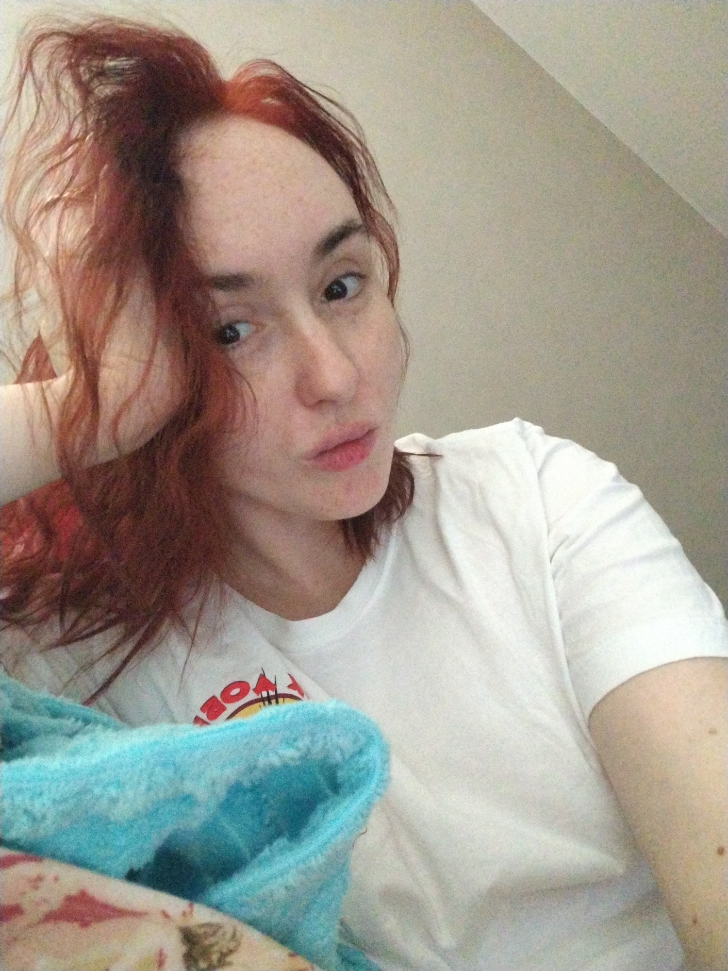 A woman with red hair rests her head on her hand, looking at the camera. She wears a white t-shirt with a graphic design, partially obscured by a light blue blanket. The background is a plain, light-colored wall. There is no indication of the location being Izhevsk, Russia, or any specific city.