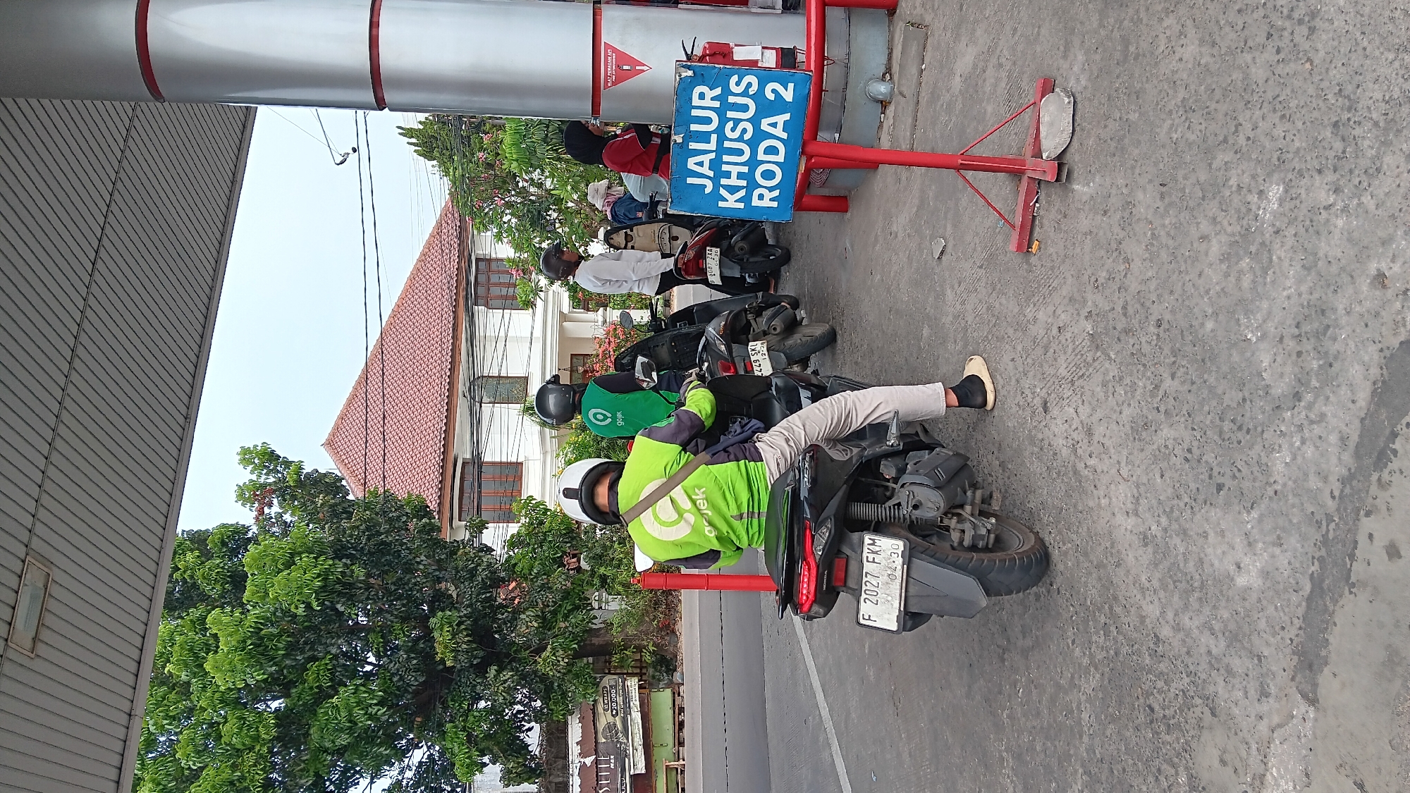 The image depicts a scene at a gas station in Depok, Indonesia. In the foreground, a person wearing a bright green Gojek jacket and a white helmet is sitting on a motorcycle. The motorcycle has a license plate reading "F 2027 FKM". The person is facing away from the camera, looking towards a row of other motorcycles and people lined up at the gas pumps.
Behind the first motorcycle, another person in a green Gojek jacket and black helmet is visible. Further back, there are several other motorcycles and individuals. To the right of the motorcycles, a blue sign with white text reads "JALUR KHUSUS RODA 2", which translates to "SPECIAL LANE FOR 2-WHEELED VEHICLES". A red fire extinguisher is mounted on a pole next to the sign.
The gas station has a covered area with a metallic ceiling, supported by a large cylindrical pillar painted with red and silver stripes. In the background, a white building with a reddish-brown tiled roof is visible, surrounded by trees and greenery. The ground is paved with concrete. The overall atmosphere suggests a typical day at a busy gas station, possibly during peak hours.