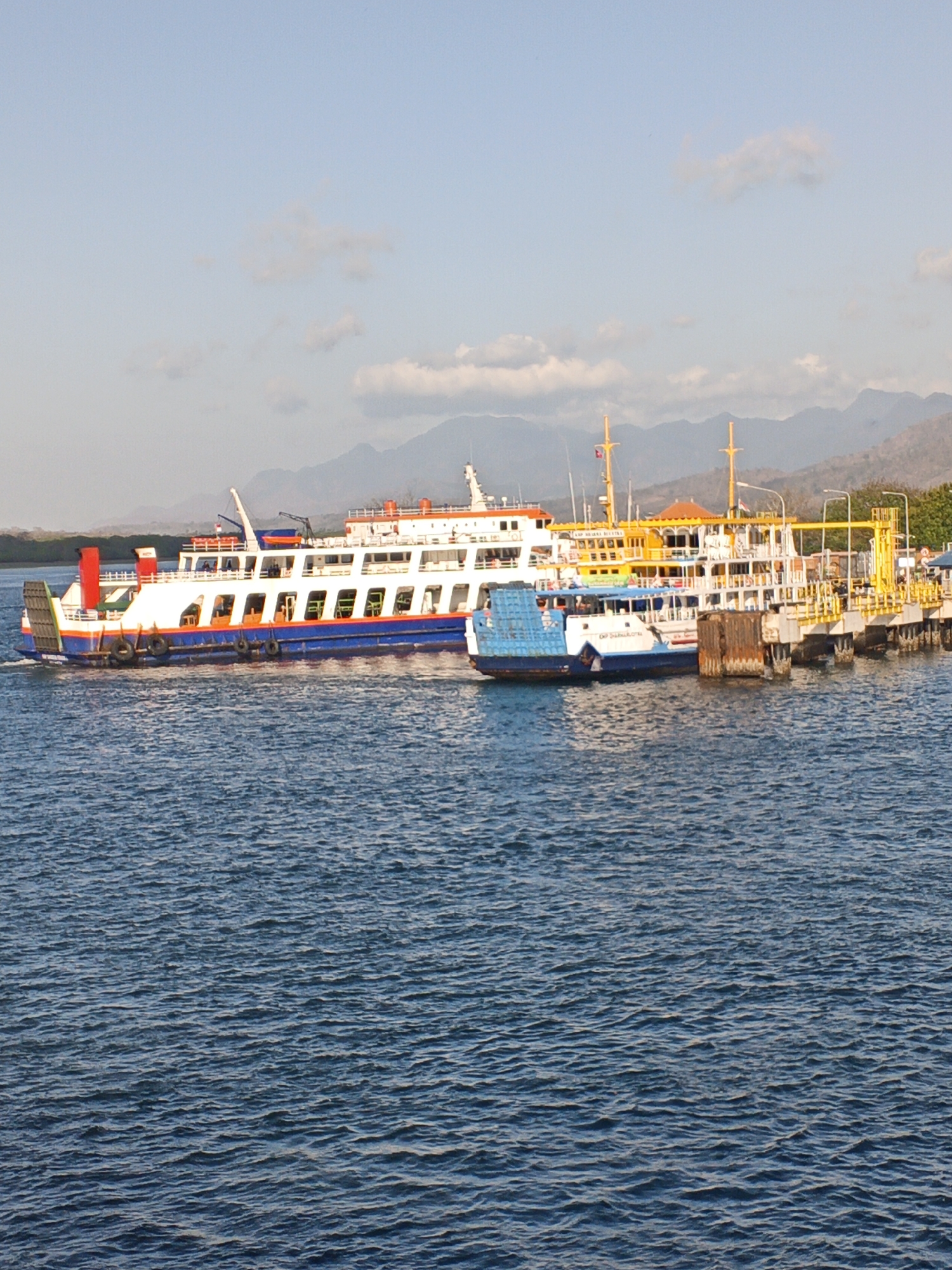 In the city of Melaya, Indonesia, a ferry terminal is depicted with multiple ferries docked or arriving. The main ferry, a large white vessel with blue and red accents and a red smokestack, is on the left. Another ferry, predominantly white with blue detailing and a blue cabin, is positioned closer to the dock on the right. The dock itself is a yellow structure with yellow railings and lights. In the background, rolling hills and mountains rise under a clear blue sky with scattered clouds. The water is calm and blue, reflecting the sky and the vessels. No people are clearly visible in the image.
