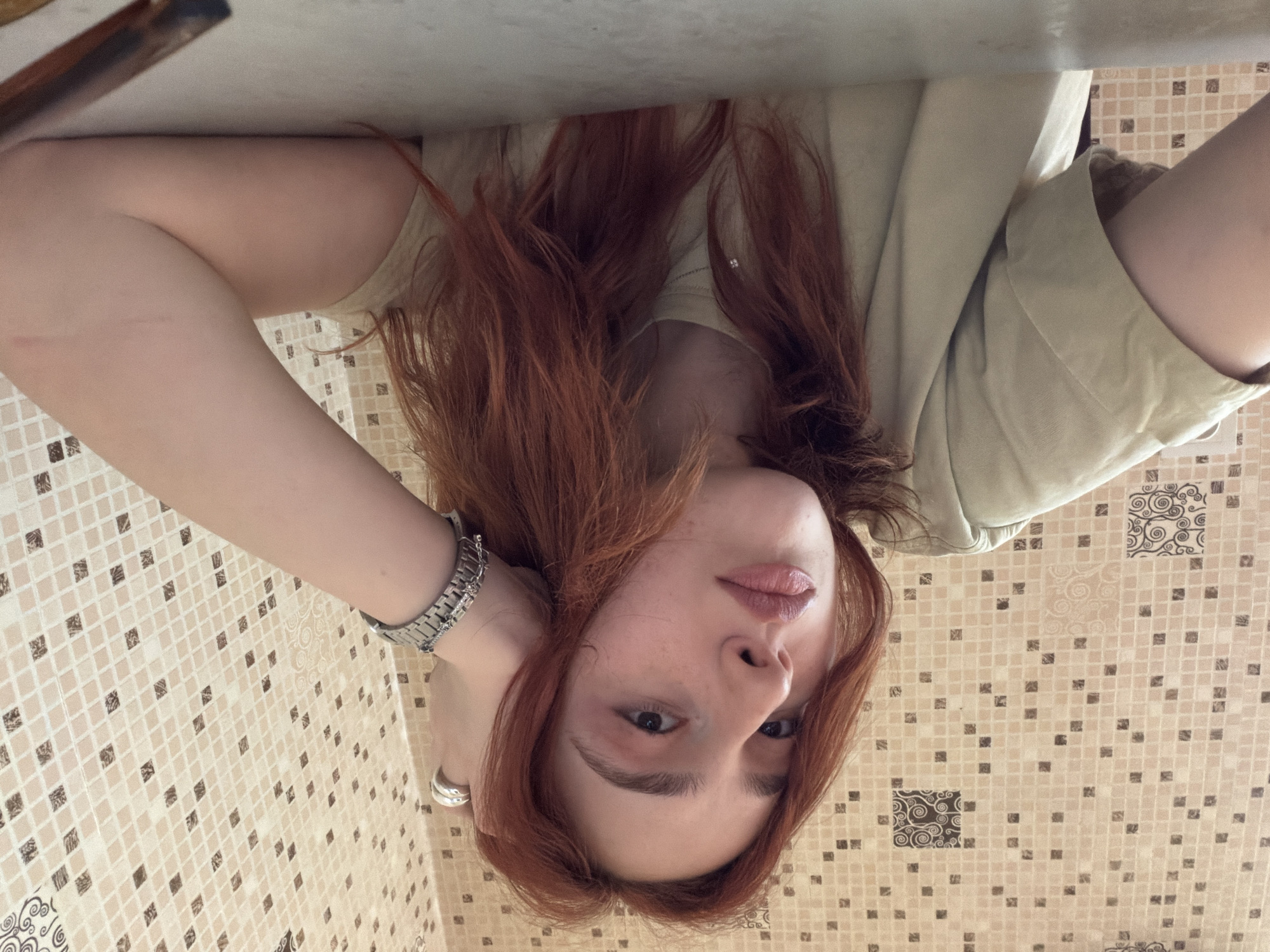 A woman with auburn hair, fair skin, and wearing a watch is posing for a selfie. She is wearing a light green top. She is positioned in what appears to be a bathroom, given the tiled wall. There are no indications that this image was taken in Tbilisi, Georgia.