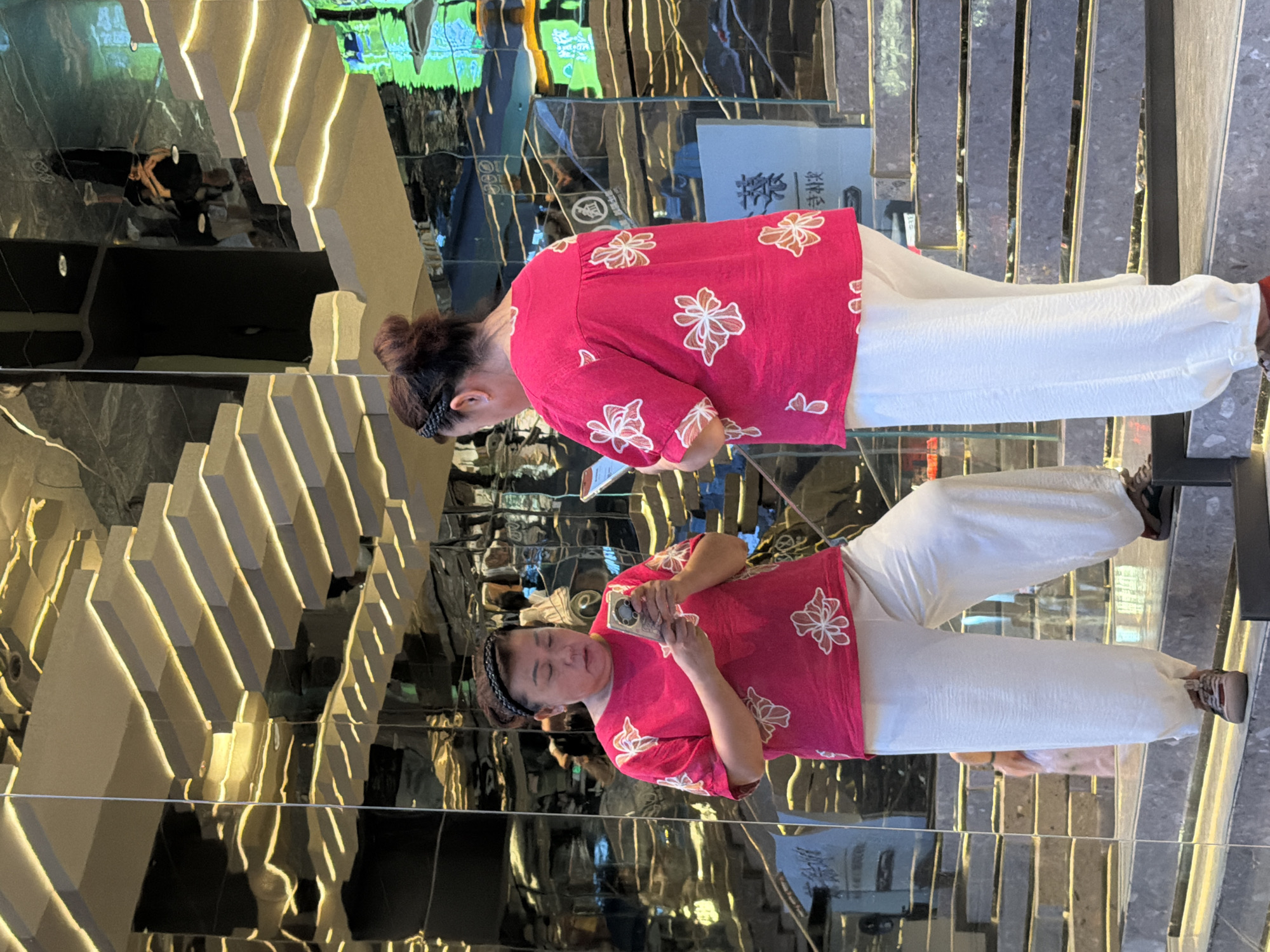 A woman with her hair pulled back in a bun is standing on a mirrored staircase in Yuzhong District, China, looking at her phone. She is wearing a bright pink shirt with white floral patterns and loose-fitting white pants. The staircase has illuminated steps and is surrounded by reflective surfaces that create a sense of depth and repetition. In the background, other people and architectural elements are visible in the reflections. The scene appears to be an indoor public space, possibly a shopping mall or a modern building.