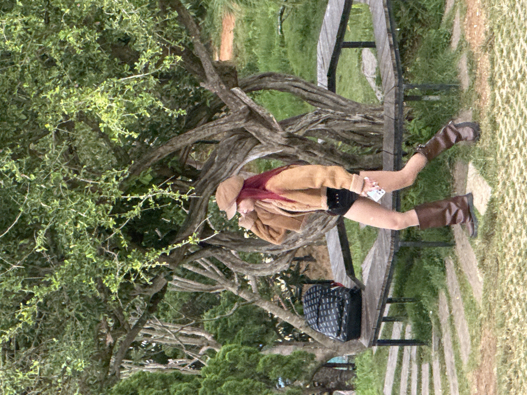 A person with long, red hair wearing a tan jacket, black shorts, and brown boots stands in front of a uniquely twisted tree trunk in Thôn 5, Vietnam. The individual is holding a white smartphone in their right hand and appears to be adjusting their tan cap with their left. A black backpack with a white patterned design rests on a wooden bench to the left of the person. The bench is situated on a paved path with a grassy area surrounding it. The background consists of lush green trees and foliage.
