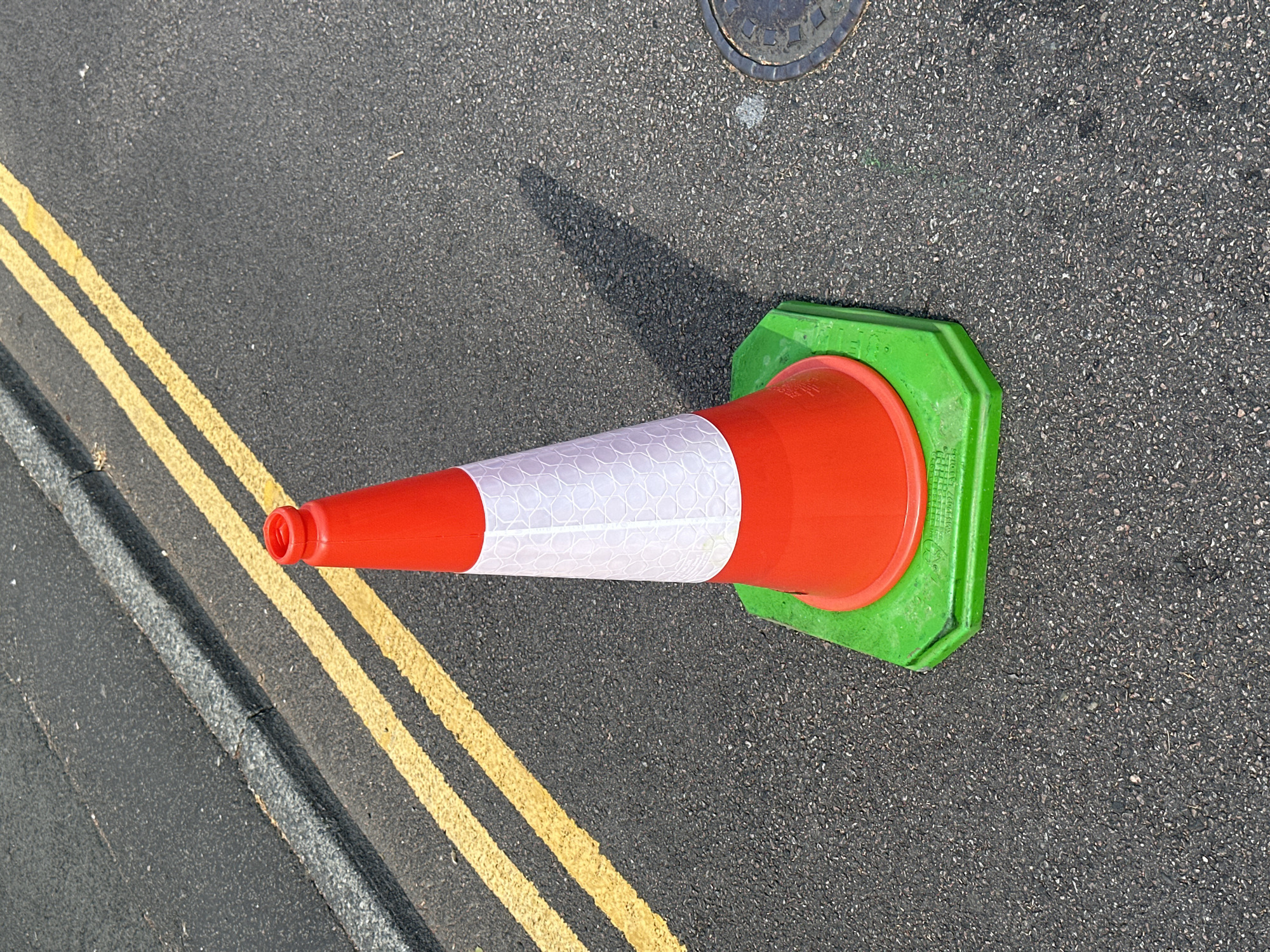 A single, bright orange traffic cone with a white reflective band stands on a street in Tunbridge Wells, United Kingdom. The cone is placed on a bright green hexagonal base. It is positioned on asphalt, with double yellow lines painted on the road to the left of the cone. The lines are slightly faded. To the far left of the image, a grey curb is visible. The cone casts a long shadow to the right. A manhole cover with a circular pattern is partially visible on the asphalt to the lower right. The scene appears to be a street, likely in a residential or commercial area, given the presence of road markings and a manhole cover. The lighting suggests it is daytime, with the sun casting a distinct shadow. There are no people or vehicles visible in the frame.