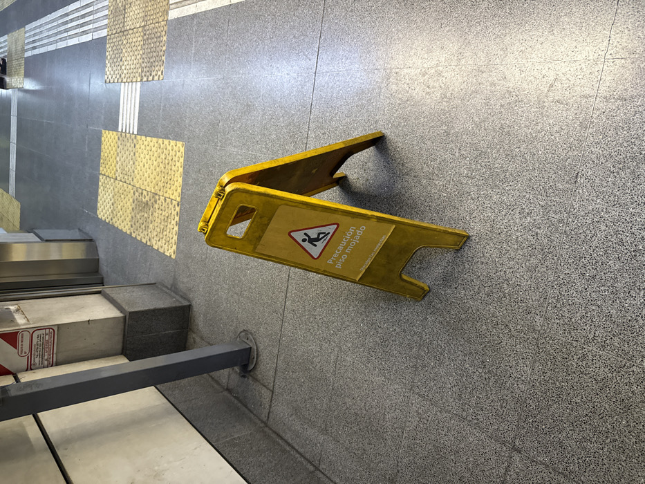 A yellow "Caution Wet Floor" sign sits on a gray-tiled floor near a set of steps. The sign includes an image of a person slipping, along with the Spanish text "Precaución piso mojado." Textured yellow safety tiles are partially visible on the floor nearby. A gray metal rail is present next to the steps. The overall scene suggests an area requiring caution due to a potential slipping hazard.