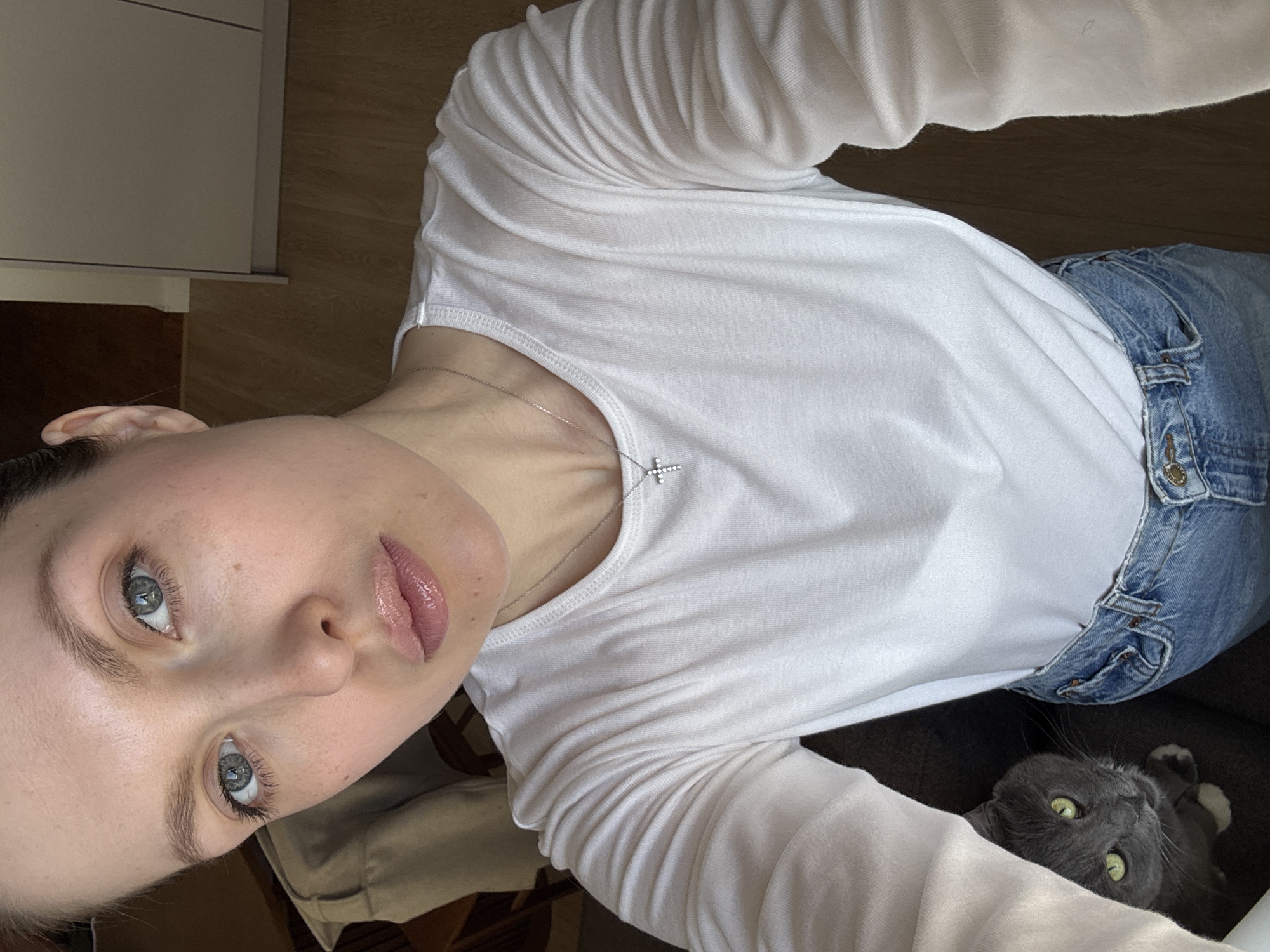 A selfie angled downward shows a woman wearing a white long-sleeved shirt and blue jeans. A silver necklace with a cross pendant is visible on her chest. A gray cat with green eyes peeks out from under her jeans. The scene appears to be indoors, with a glimpse of a wooden floor and cabinet in the background. There is no information available to determine if the image was taken in Moscow, Russia.