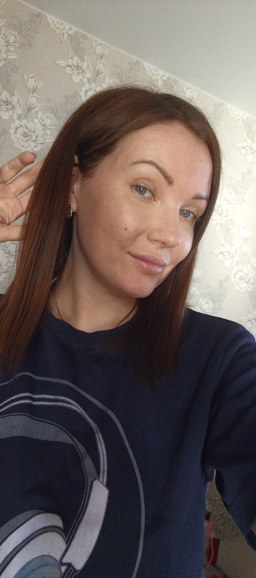 A woman with shoulder-length auburn hair, blue eyes, and fair skin is looking at the camera in an indoor setting. She is wearing a navy blue long-sleeved shirt with a graphic of headphones on the front. The woman is holding her left hand up near her face, with her fingers slightly splayed. She has a gold chain necklace and simple gold earrings. The background is a wall covered in patterned wallpaper. The location is Chaikovsky, Russia.