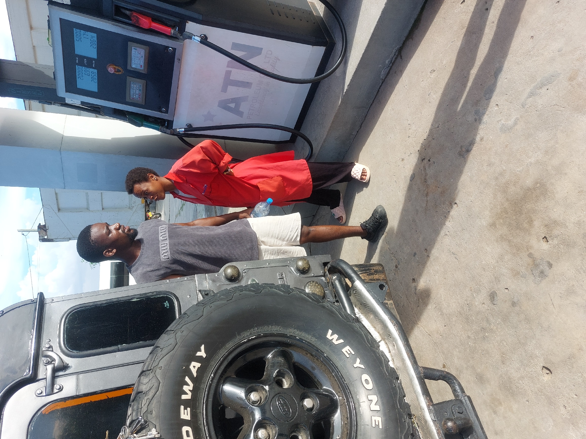 A man in a grey t-shirt and white shorts is standing near a grey vehicle, specifically a Land Rover Defender, at a petrol station. The vehicle has a spare tire mounted on the rear. A person wearing a red jacket is standing next to the man. They are near a petrol pump which indicates 20,000 on the sale and 2,858 as the price. The petrol station has the brand "ATN Petroleum Ltd". The location appears to be in Dar es-Salaam, Tanzania based on the brand.