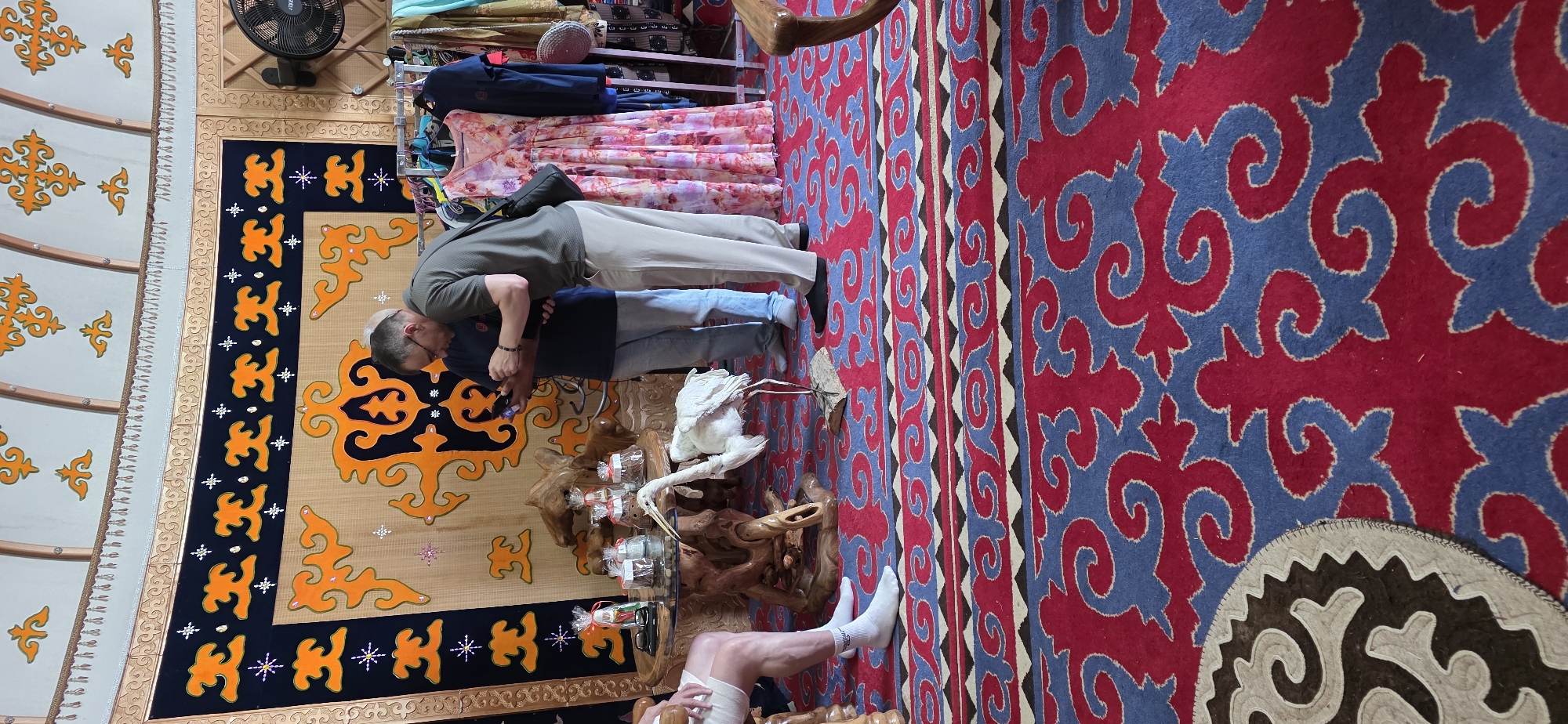 The image depicts the interior of a yurt, likely located in Bulan-Segettu, Kyrgyzstan, given the context. In the center, two men are standing, engrossed in their phones. One man, wearing a green polo shirt and beige pants, leans forward, holding his phone. The other man, in a navy blue shirt and light blue jeans, stands beside him, also looking at his device. Between them and slightly to the left, a stuffed bird, resembling a stork or heron, stands on a wooden base. In front of the men is a round, wooden table with intricate carvings, on which several wrapped items, possibly gifts or food, are placed. To the right, a rack holds various colorful garments, including a floral-patterned dress. The yurt's interior features decorative wall hangings with orange and black patterns and a carpet with a striking red and blue design. The ceiling of the yurt is visible at the top, adorned with painted motifs. A person's legs, wearing white socks, are visible on the far left, suggesting another individual is present but mostly out of frame. The overall impression is of a cultural setting, possibly a shop or accommodation, where traditional elements blend with modern technology.