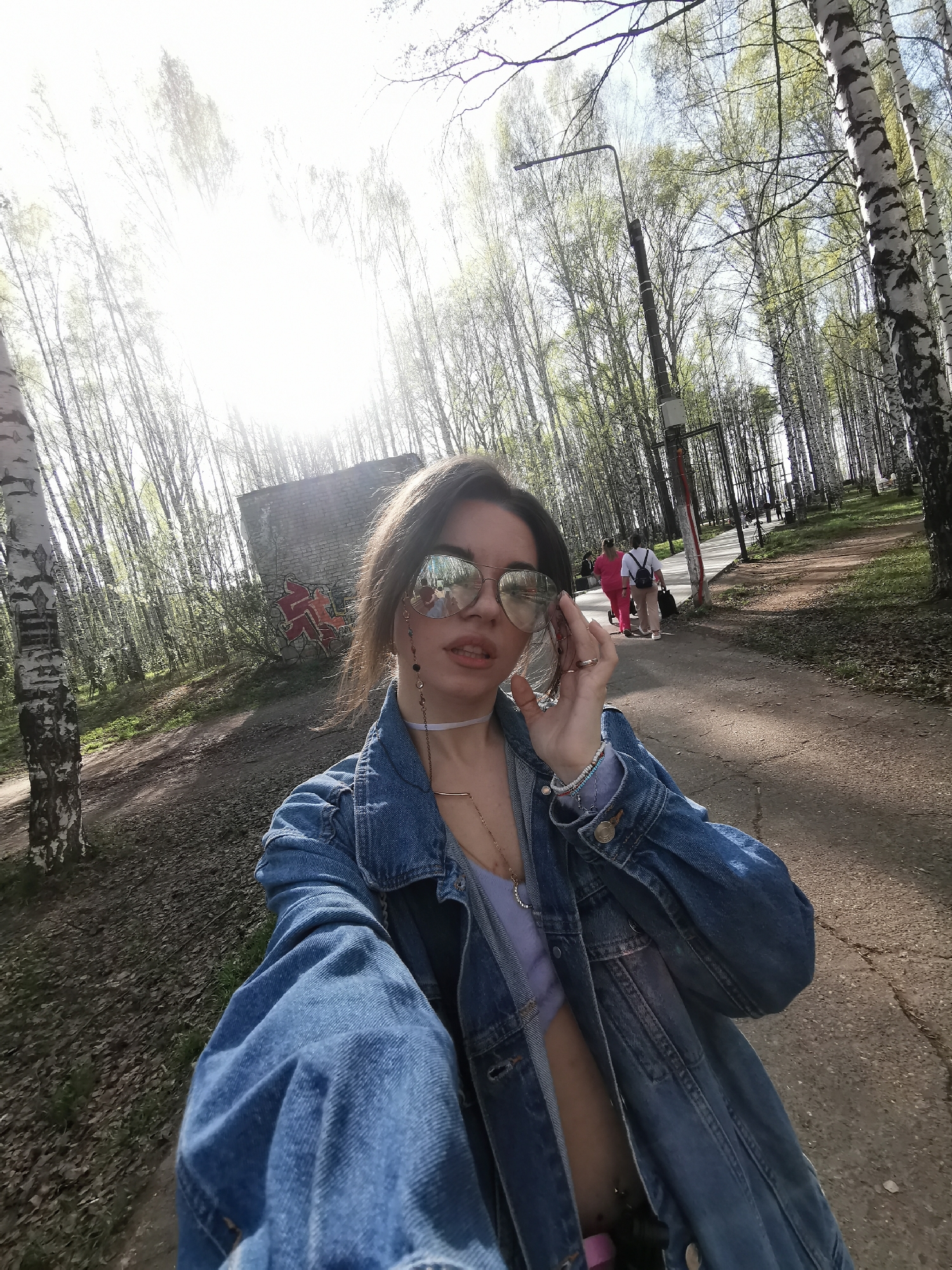 A person is taking a selfie in a park. The person has brown hair and is wearing sunglasses, a denim jacket, and a light purple top. They are holding the sunglasses with their right hand. In the background, there is a paved path, birch trees, and a building with graffiti on it. Further down the path, several people are walking. There is also a street light visible. The sun is bright, casting light through the trees. The location is in a park, possibly in the city of Unknown, Unknown.