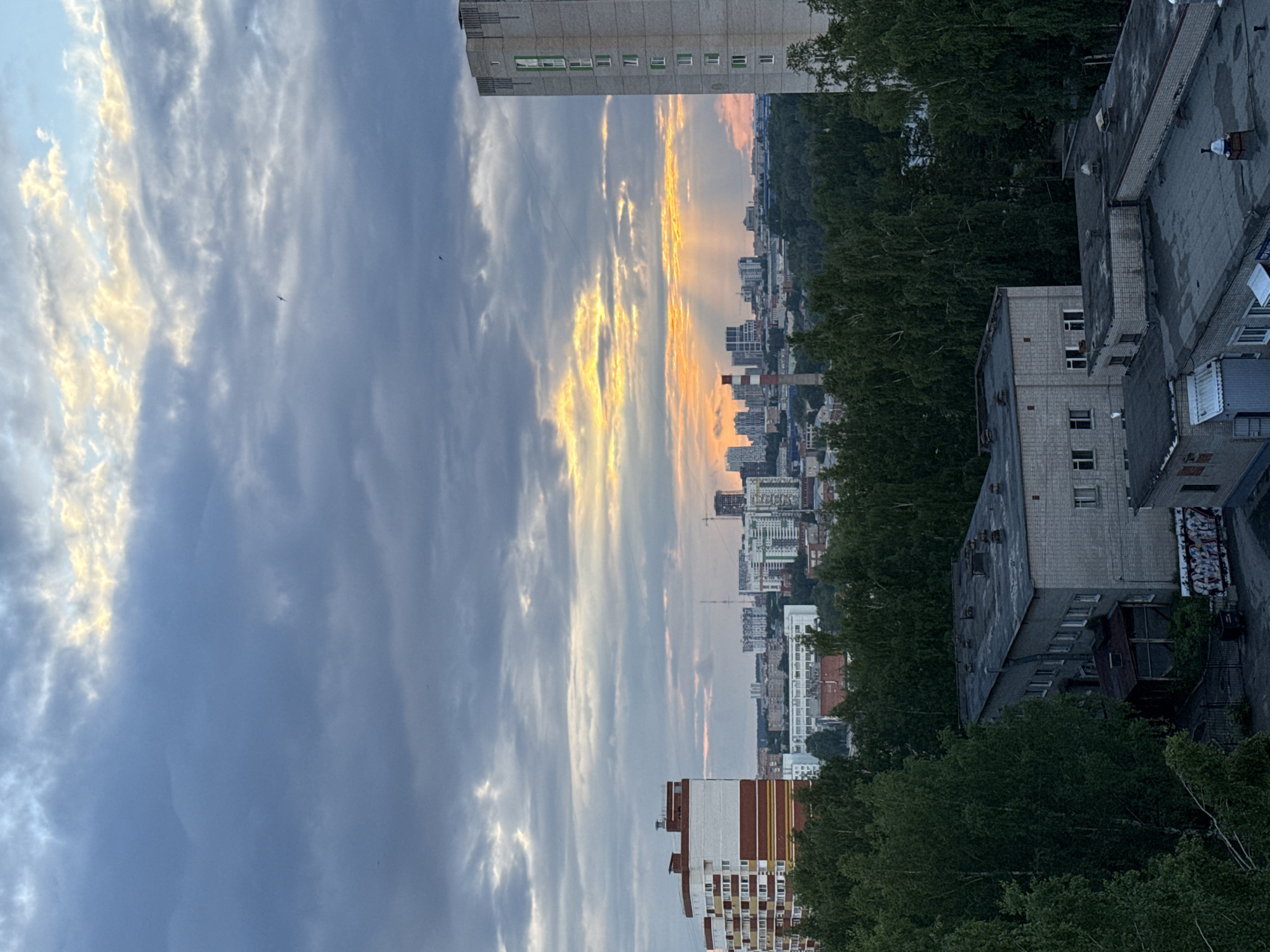 This image, correctly oriented horizontally, captures a panoramic view of an urban landscape in Izhevsk, Russia, at either sunset or sunrise. The sky dominates the upper portion, displaying a dramatic contrast. On the left, patches of clear blue peek through scattered white clouds, while towards the right, the clouds are intensely illuminated with warm golden and orange light, indicative of the sun's low position. Several small, dark specks, likely birds, are visible against the sky.
The lower half of the image reveals a sprawling city. In the foreground and mid-ground, a mix of multi-story buildings with flat roofs, characterized by their grey, blocky architecture, are prominent. These structures are interspersed with abundant, lush green trees, suggesting parks or natural areas woven into the city. One building in the lower left has distinctive red and white vertical stripes. Closer to the right, some buildings appear older and weathered; one features a patch of abstract, illegible graffiti on its side. A small, white, possibly spherical object is visible on one of the nearby rooftops.
Further into the distance, the cityscape stretches extensively, featuring numerous residential blocks and commercial buildings. A tall red and white smokestack rises above the mid-ground structures. The distant horizon shows a hazy outline of more buildings blending into what appear to be forested hills. No specific human activity is discernible, presenting a serene yet expansive view of the city.