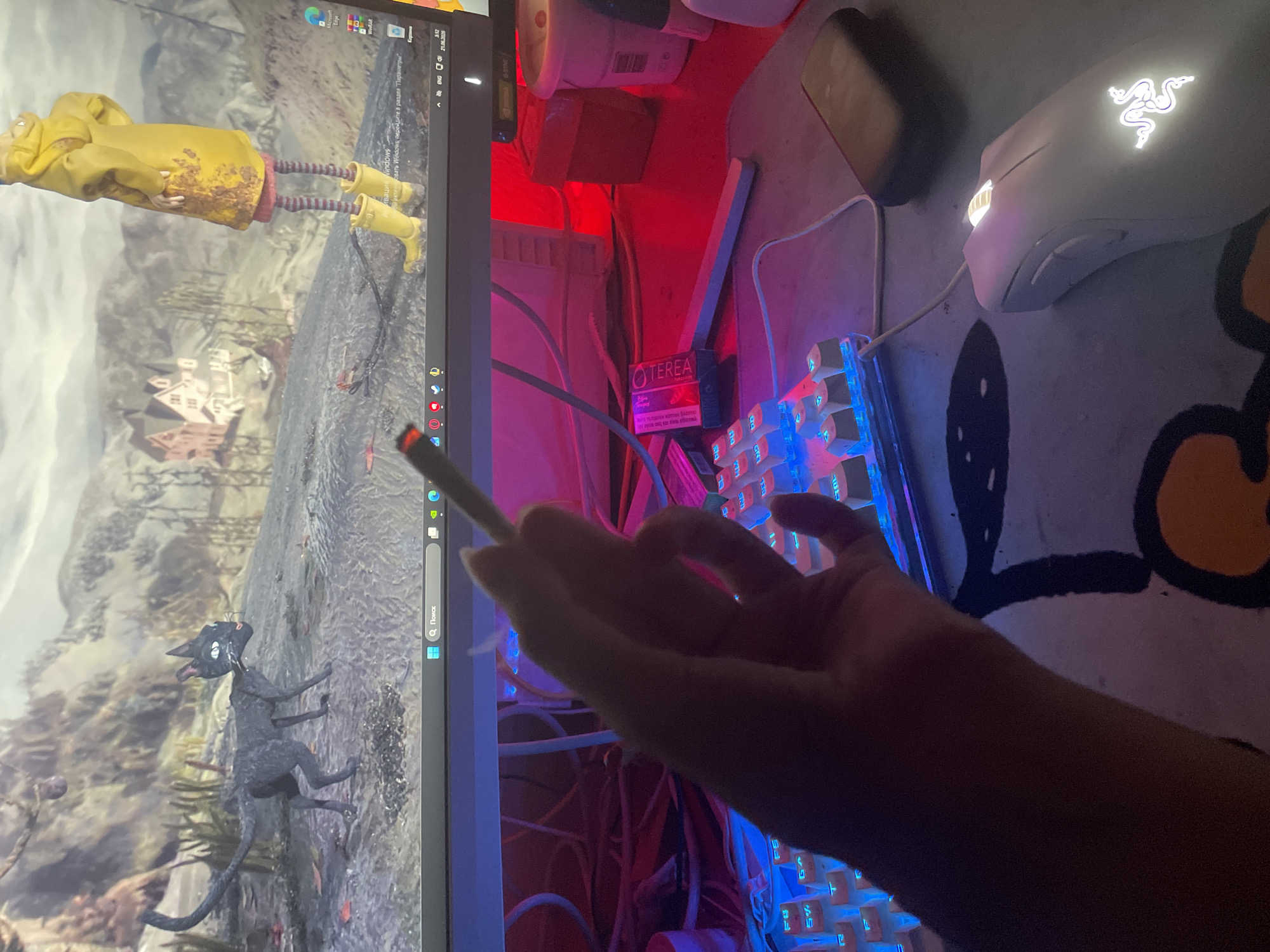The image shows a person holding a lit cigarette in their right hand, with the cigarette smoke visible. The person's hand is in the foreground, partially obscuring a white keyboard with blue backlighting. In the background, a computer monitor displays a scene from the movie "Coraline," featuring a black cat and a doll-like character in a yellow raincoat. The desk is illuminated by red lighting. To the right of the keyboard, a white computer mouse with a glowing logo is visible on a patterned mousepad. Several cables are visible behind the keyboard. There is no indication of the location being in Neoi Epivates, Greece.