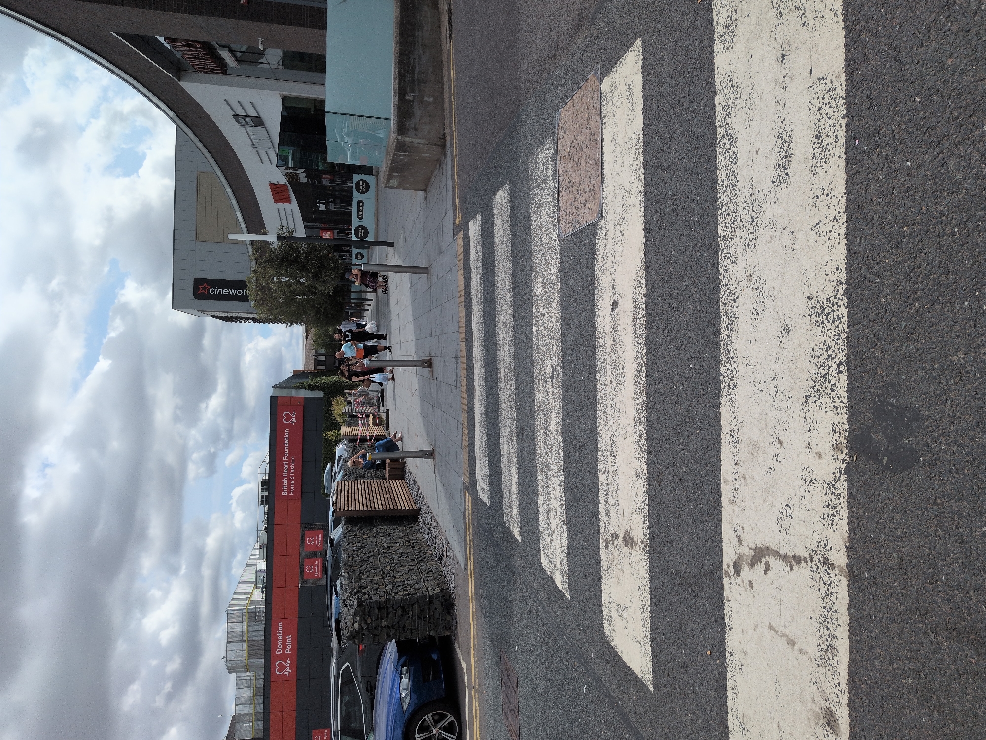 The image shows a pedestrian crossing in front of a row of shops in Telford, United Kingdom. On the left, a blue car is parked next to a stone wall. In the background, people are walking on the sidewalk in front of buildings that include a Cineworld cinema and various retail stores. The sky is cloudy.