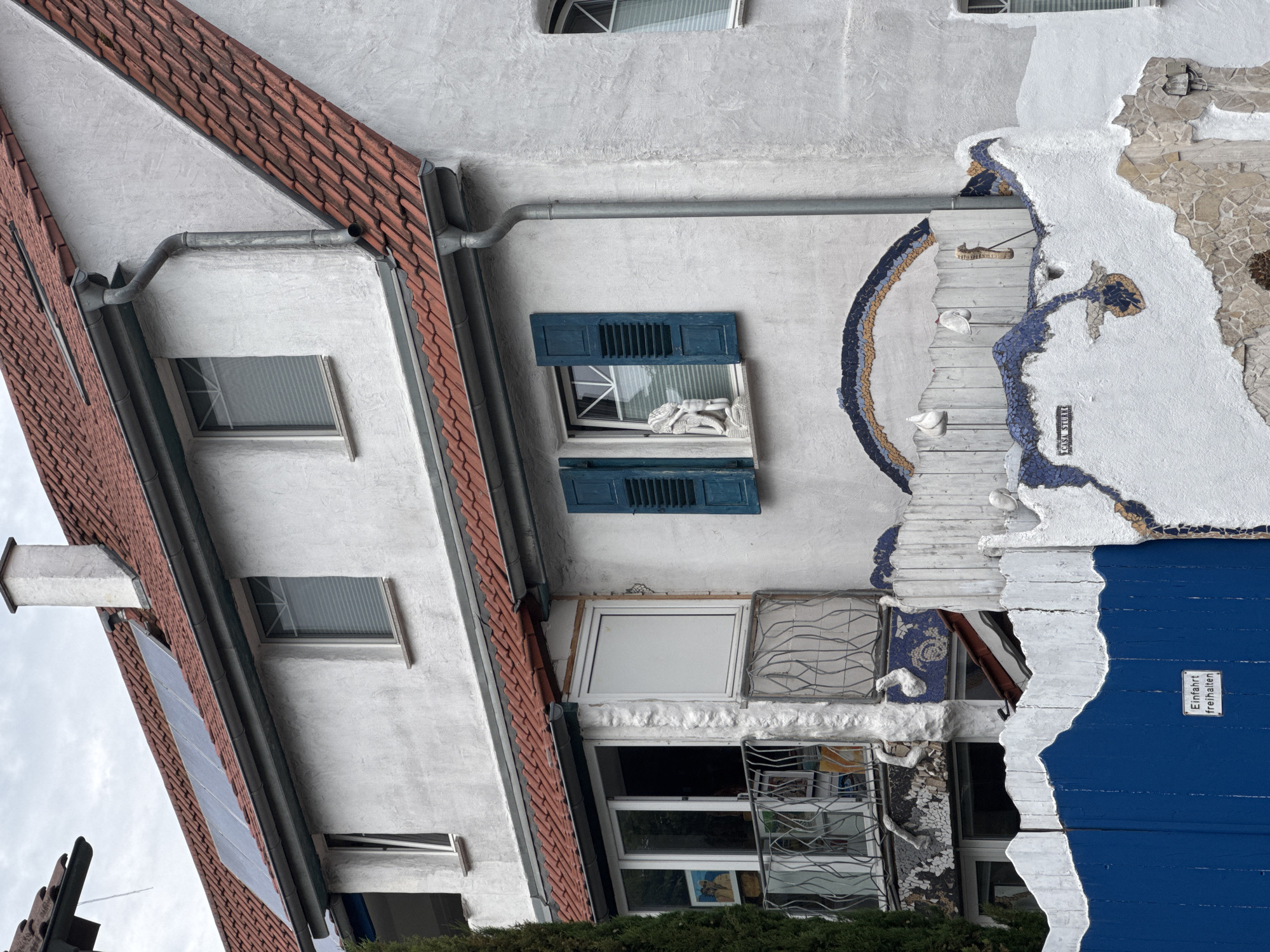 The image shows a white, two-story house with a red-tiled roof and solar panels. There are several windows with blue shutters and blinds. A balcony with decorative metal railings extends from the upper floor. The house is decorated with colorful mosaics and sculptures, including a seahorse and two white birds. A bright blue wooden fence is in the foreground, with a sign that reads "Einfahrt freihalten" (Keep driveway clear). The house is located in Bietigheim-Bissingen, Germany, a town situated on the Enz river. The sky is overcast.