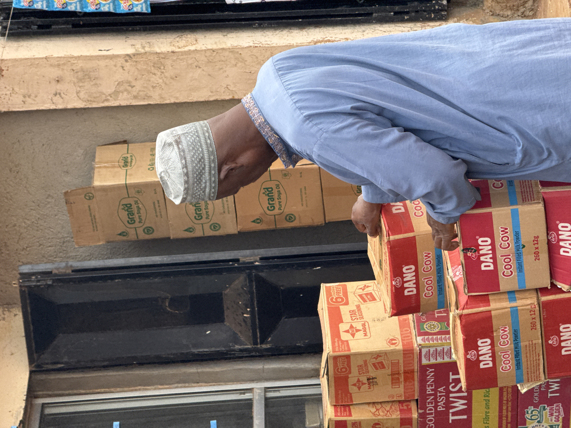 A person, possibly a man, wearing a light blue robe and a patterned white cap is bent over, looking at a stack of boxed goods. The boxes are predominantly red and brown, with visible text indicating brands such as "DANO Cool Cow," "Grand Pure Soya Oil," and "MAGGI STAR SEASONING." The boxes are stacked high, suggesting a commercial setting, possibly a market or a shop. Some of the boxes are open, revealing the contents within. The scene appears to be outdoors, with a rough, textured wall in the background. The lighting suggests it could be daytime. The overall impression is that of someone engaged in the business of stocking, arranging, or inspecting merchandise.