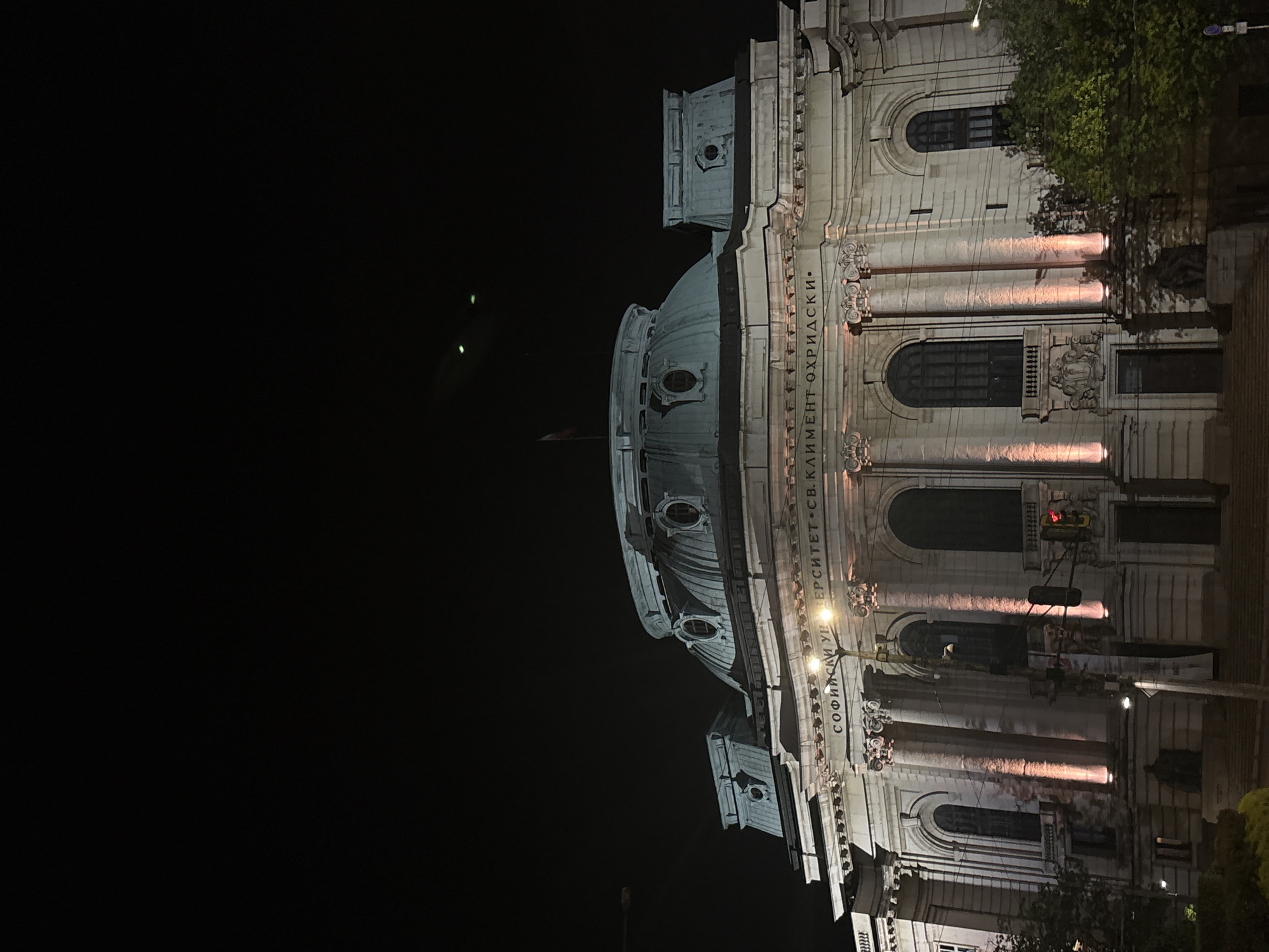 The image depicts Sofia University St. Kliment Ohridski in Sofia, Bulgaria at night. The building's facade is illuminated, showcasing its dome, columns, and architectural details. The inscription "СОФИЙСКИ УНИВЕРСИТЕТ СВ. КЛИМЕНТ ОХРИДСКИ." is visible. There is a traffic light, a flag, and trees in the background. The sky is dark.