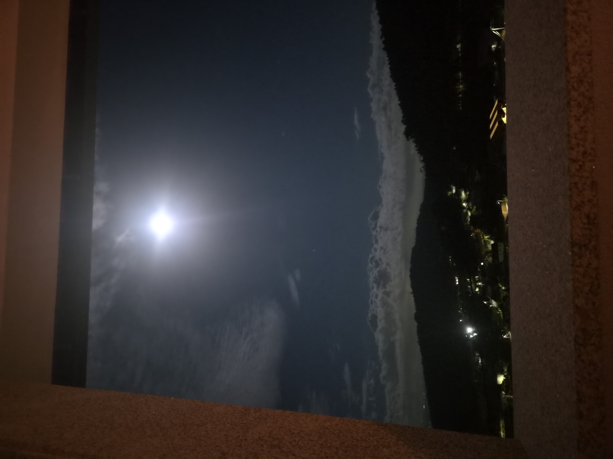 The image shows a nocturnal, panoramic view through an opening framed by a textured, dark brown material on the left and a similar material on the bottom. The sky is dark blue with a bright, luminous full moon positioned slightly left of center, emitting a strong glow. Wispy clouds are scattered across the sky, some appearing to be illuminated by the moonlight. Below the sky, a line of dark, silhouetted hills is visible, dotted with faint lights from what appears to be a town or village. These lights are predominantly yellow and white, suggesting artificial illumination from houses and streetlights. The terrain is uneven, with slopes and vegetation visible on the hillsides. In the foreground, below the moonlit sky and behind the bottom frame, there are structures, possibly outdoor seating or awnings, illuminated by artificial lights. The overall impression is a view of a rural or suburban area at night, under a bright moon. The image does not contain any identifiable people or specific landmarks that would allow for pinpointing a precise location within Barcelos, Portugal, but it depicts a scene consistent with a hilly or mountainous region at night.