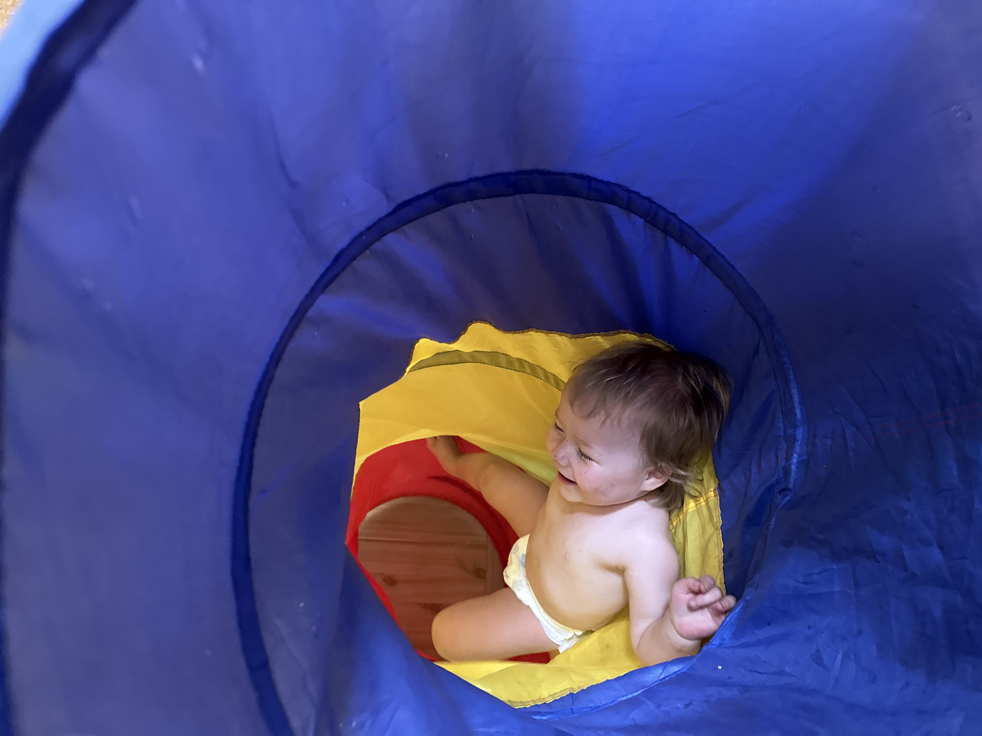 A baby in a diaper is inside a play tunnel with blue, yellow, and red sections. The baby appears to be looking towards the red opening of the tunnel. The floor is visible through the red opening.