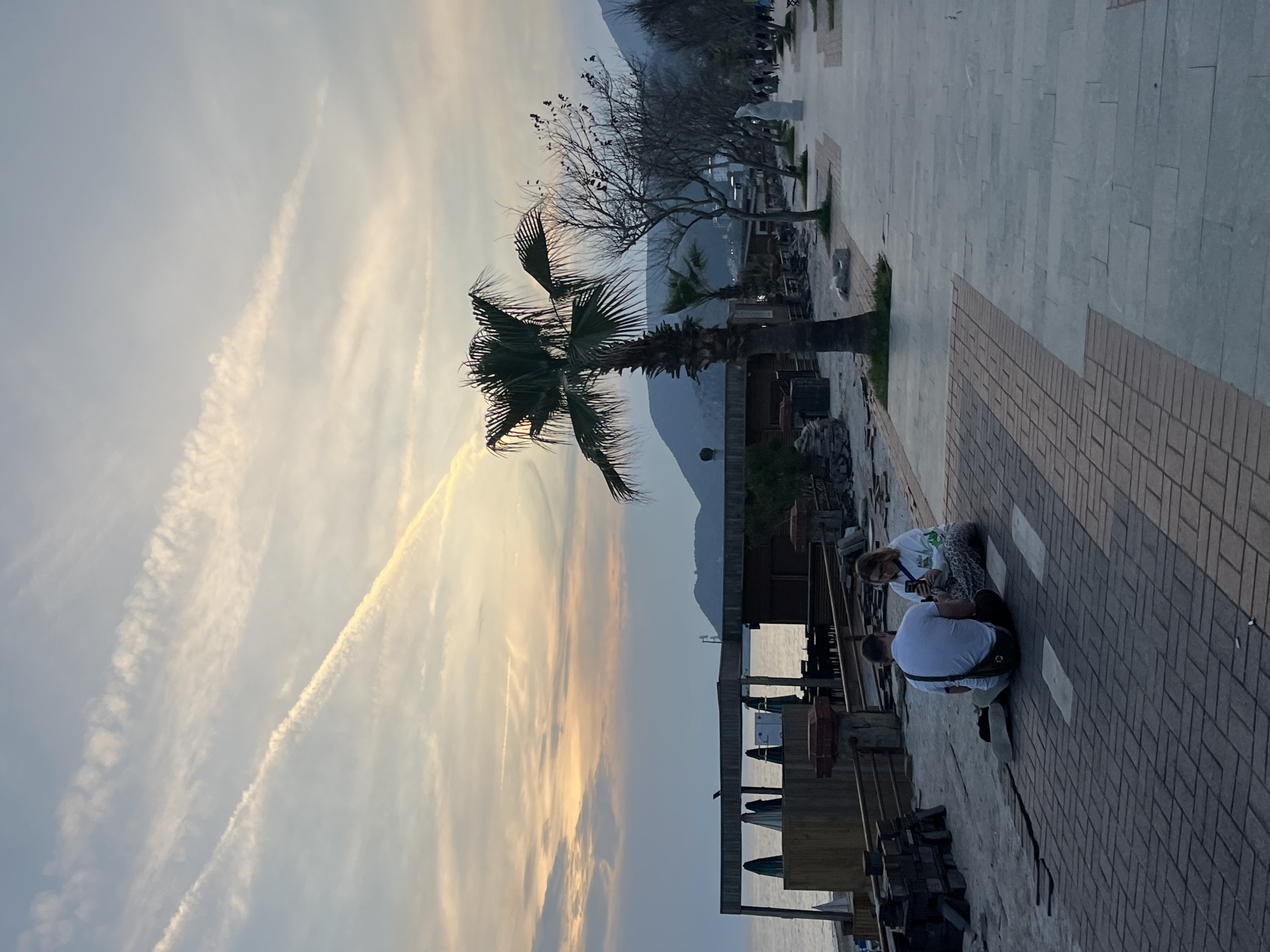 The image captures a tranquil scene at sunset in Alanya, Turkey. A prominent palm tree stands to the left, silhouetted against the sky. Beyond the tree, the horizon is marked by mountains.
In the foreground, on a tiled pathway, two individuals are seated. One is wearing a white shirt, while the other is in a patterned shirt. They appear to be focused on a mobile device.
Behind them, a partially constructed structure with wooden supports and green umbrellas suggests an outdoor seating area, likely part of a restaurant or cafe. The sky displays contrails.