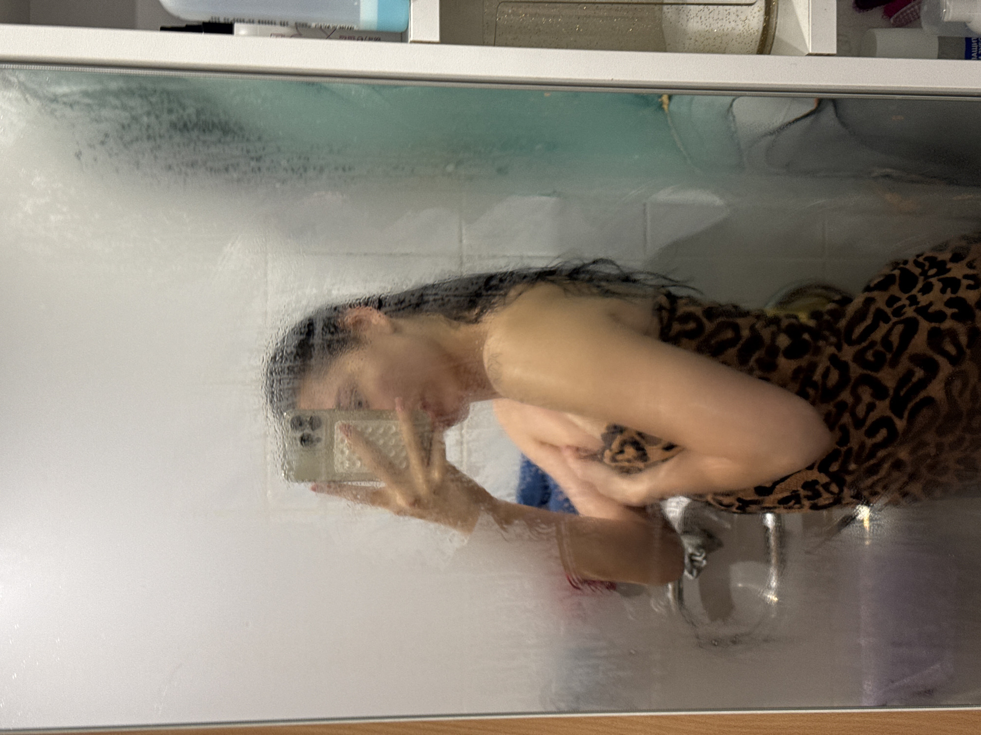 A young woman, dressed in a leopard print towel, takes a selfie in a steamy bathroom mirror. Her dark hair is wet and hangs down her back. She is holding her phone in her right hand, angled to capture her reflection. Her left arm is bent across her chest, obscuring part of her body. The mirror is covered in condensation, and the background is blurred. To the right of the mirror, a white shelf holds various toiletries. The overall impression is that of a candid, personal moment in a private space. The image is set in a bathroom in the city of Chaikovsky, Russia.