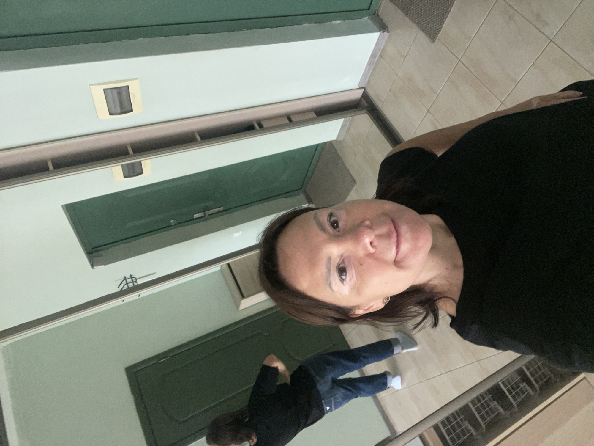 A medium shot captures a woman in a black shirt, with a person in a black shirt and blue jeans reflected behind her. The reflection suggests they are standing in a corridor with green doors and light green walls. The floor is tiled in light brown. A motion detector is visible on the wall. The location is unspecified, and there is no indication of Sochi, Russia.