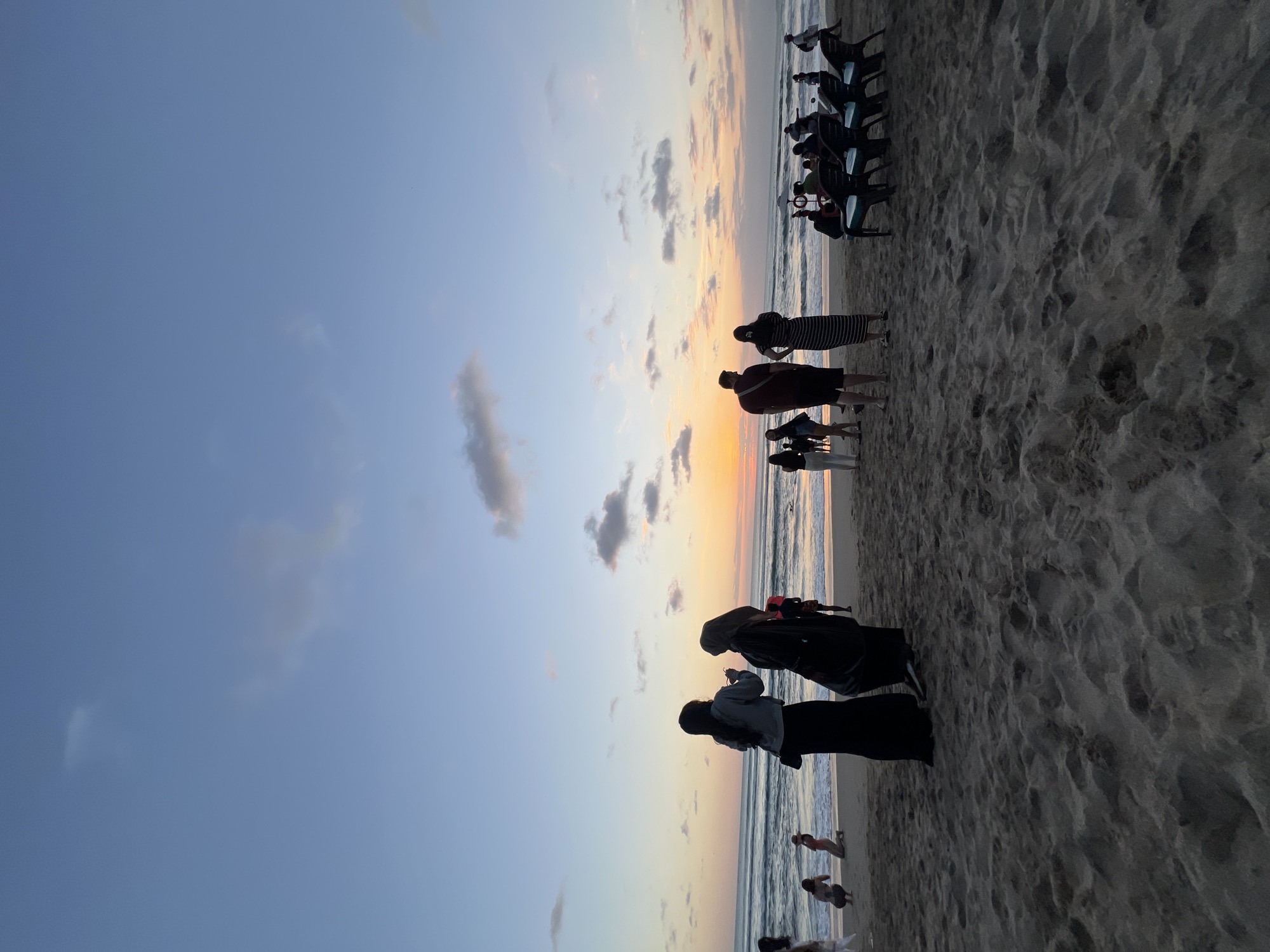 A group of people are on a sandy beach in Kuta, Indonesia, during sunset. The sky is a gradient of blue and orange, with scattered clouds. The ocean is calm with gentle waves. Some people are standing and looking at the sunset, while others are sitting on chairs arranged in rows. There are also people walking along the shoreline and some individuals in the water. The sand is dark and textured, with footprints visible. The overall atmosphere appears relaxed and serene.
