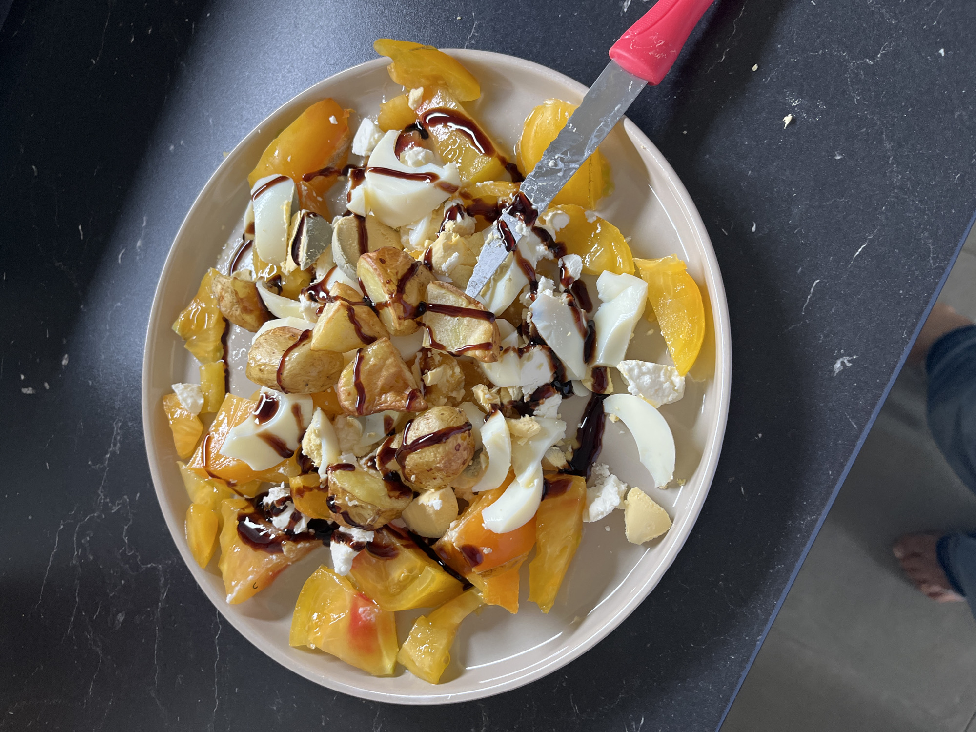 A plate of chopped yellow tomatoes, sliced hard-boiled eggs, crumbled white cheese, and roasted potato pieces is drizzled with a dark sauce. A knife with a pink handle is positioned on the right side of the plate, with its blade cutting into the food. The plate rests on a dark, marbled countertop. The scene is indoors, likely a kitchen. There are no discernible landmarks or features indicating the specific location in Chambœuf, France. The image is a close-up shot of the food and plate.