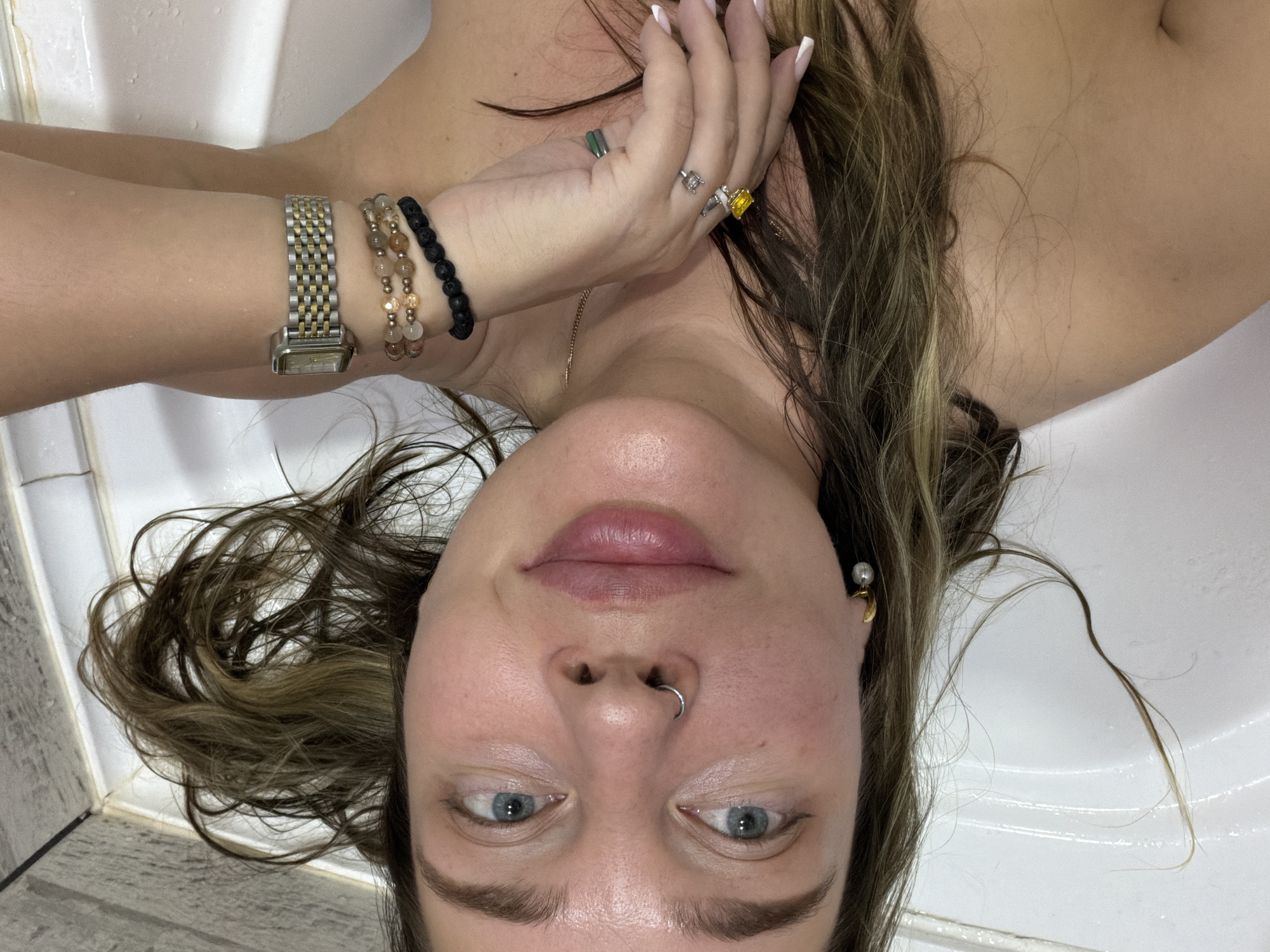 A fair-skinned woman is reclining in a white bathtub. Her face is centered and angled towards the viewer. She has light blue eyes, prominent eyebrows, a silver nose ring, and pink lips. Her hair is a mix of brown and blonde highlights. She is wearing a gold earring with a pearl-like accent on her right ear. Her left wrist is adorned with a gold and silver watch and two beaded bracelets, one with brown and gold beads and the other with black lava-like beads. Her left hand is raised, with fingers displaying white-tipped nails and several rings, including a green stone, a silver one, and a yellow stone. The surrounding environment consists of a white bathtub and a glimpse of tiled walls in the upper left corner. The image provides no information to determine if the location is Chaikovsky, Russia.
