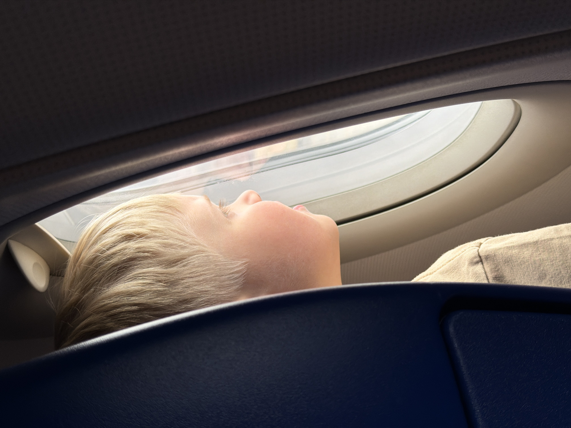 A child with fair skin and blond hair is seen from a low angle, seemingly lying or resting their head against the back of an airplane seat. The child is positioned near a plane window, appearing to look outside, though what is visible through the window is not clear. The interior of the plane, including a dark blue seat and the window panel, are visible. The location is unknown.