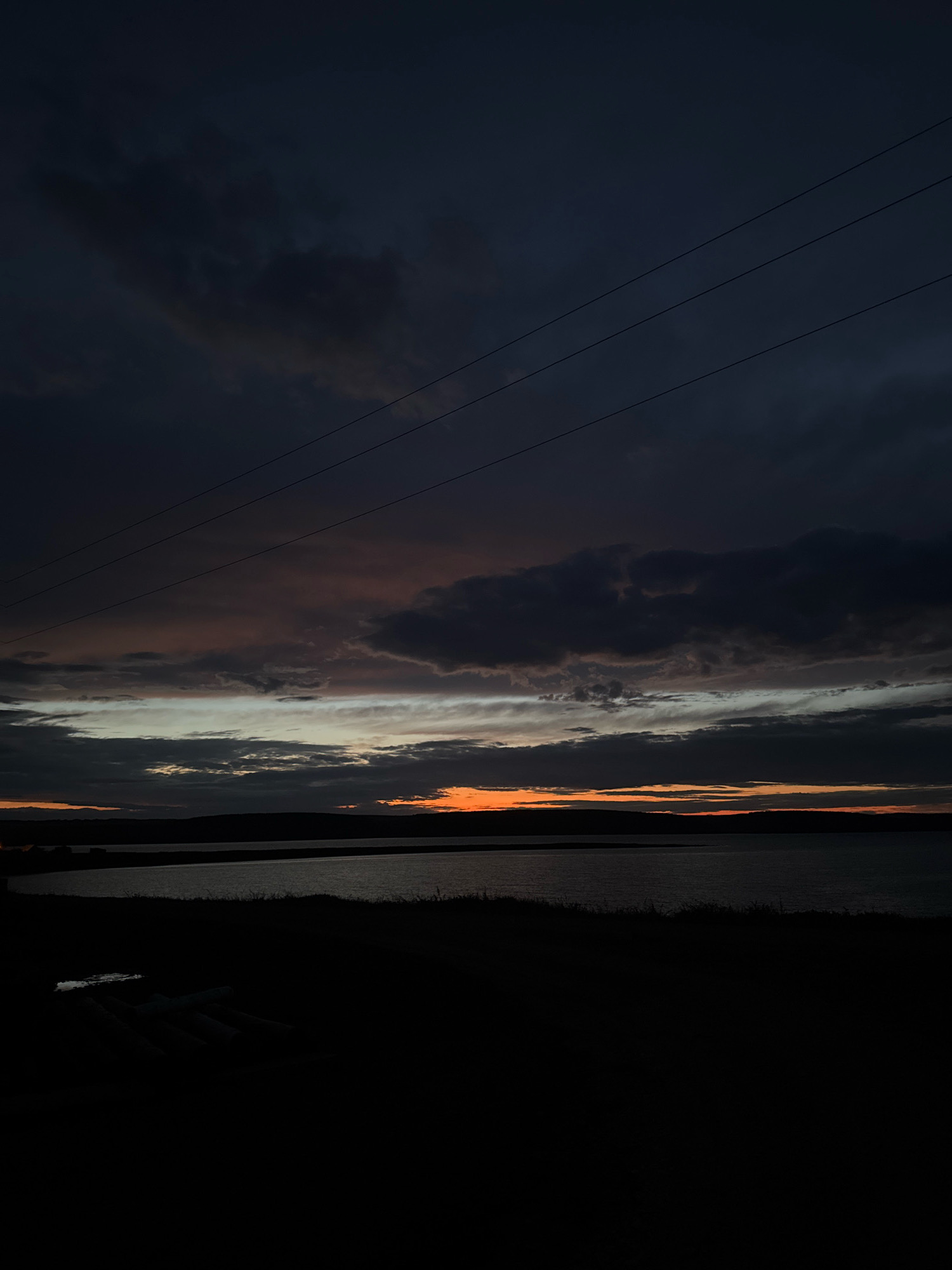 The image depicts a dark, cloudy sky over a body of water and a silhouetted landscape, likely taken at dusk or dawn. The sky is filled with heavy, dark clouds that are tinged with faint orange and pink hues near the horizon, suggesting the last remnants of sunlight. Three thin power lines are visible stretching diagonally across the upper portion of the frame. The foreground is a dark, indistinct silhouette of land, with what appears to be some light-colored objects scattered on it, possibly debris or equipment. The water in the middle ground is calm and reflects the dim light from the sky. The horizon line is low, separating the dark landscape from the dramatic sky. The overall mood is somber and atmospheric. No people or specific landmarks are identifiable in the image, and without further context, it is impossible to definitively confirm the location as Kamenka, Russia.