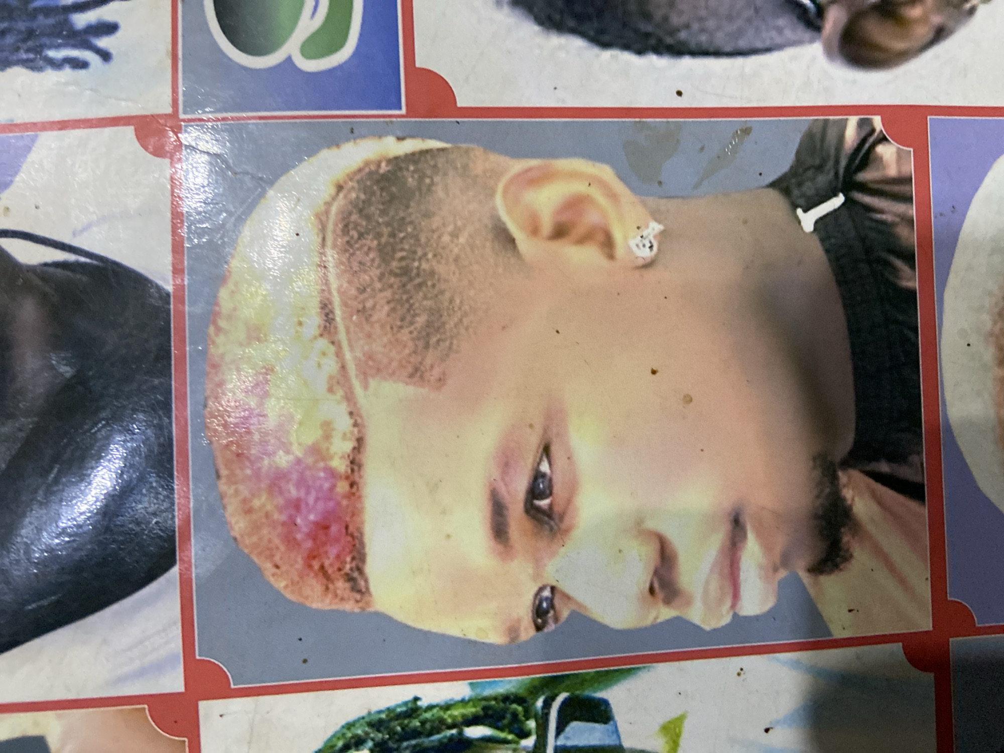 The image displays a close-up of a man's head and shoulders, featuring a stylish haircut with faded sides and a pinkish-red tint on top. He is wearing a black garment and a small, silver earring shaped like a skull on his left ear. The photograph is likely a display board from a barber shop in Kuson, Nigeria, showcasing hairstyles. The background is a collage of other images, suggesting a commercial setting. The image quality is somewhat degraded due to printing and environmental factors, with visible specks and color distortion.