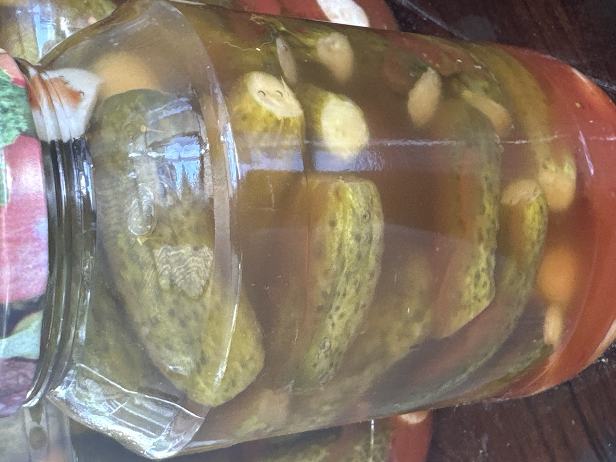 A close-up, eye-level shot shows a glass jar filled with pickles and brine, likely preserved food. The jar is clear, allowing a view of the pickles inside, some of which appear to be sliced. The brine is a reddish-brown color. The jar has a patterned lid, with visible floral designs in shades of pink and green. The lighting is somewhat dim, creating reflections on the glass. The background is blurred and dark, suggesting an indoor setting. There are no people or animals visible in the image. The image does not provide any information about its location in Yessentuki, Russia.
