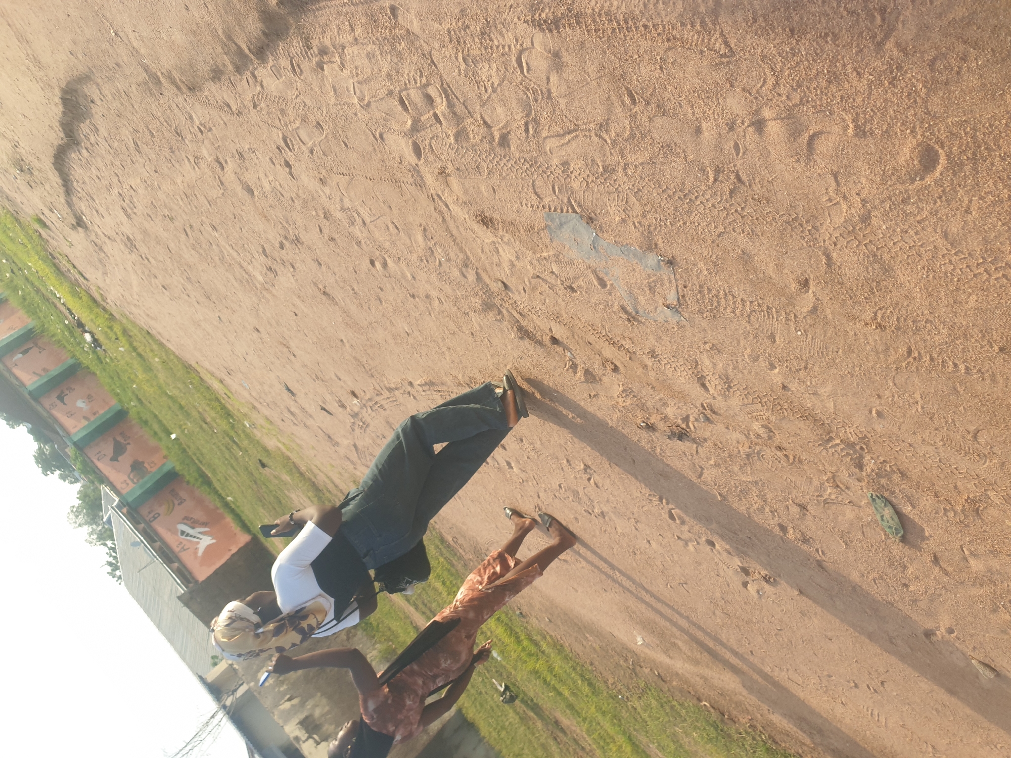 Two individuals are walking on a dirt path in Jalingo, Nigeria. The person in the foreground, facing away from the camera and to the left, is wearing a light-colored headscarf with a floral pattern, a white t-shirt, and dark jeans. They are holding a mobile phone in their right hand and appear to be taking a photo. Beside them and slightly ahead, another person in a patterned dress is walking away from the camera. The ground is sandy with visible footprints and tire tracks. In the background, a building with colorful murals and green grass is visible. The scene is illuminated by natural sunlight, casting shadows on the path.