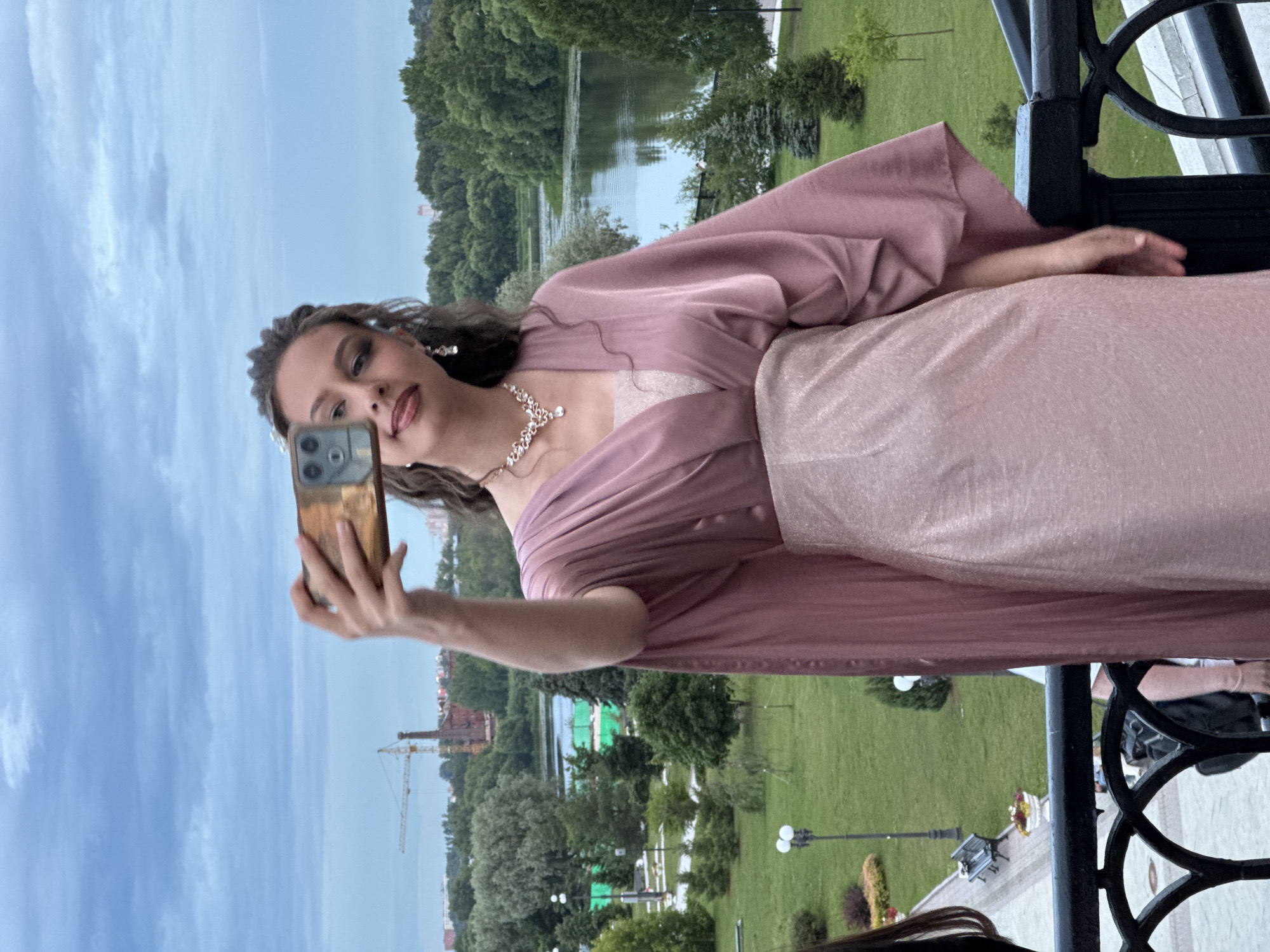A young woman with dark hair is taking a selfie with her smartphone while standing on a balcony overlooking a park with a body of water and trees. She is wearing a shimmering rose-gold dress and a diamond necklace. The sky is cloudy, suggesting it could be late afternoon or early evening. The background shows a green park, a lake, and some buildings in the distance, including a construction crane. The woman's expression is playful as she poses for the photo. The setting appears to be a scenic outdoor location, possibly during a special event given her attire. The image is taken in portrait orientation.