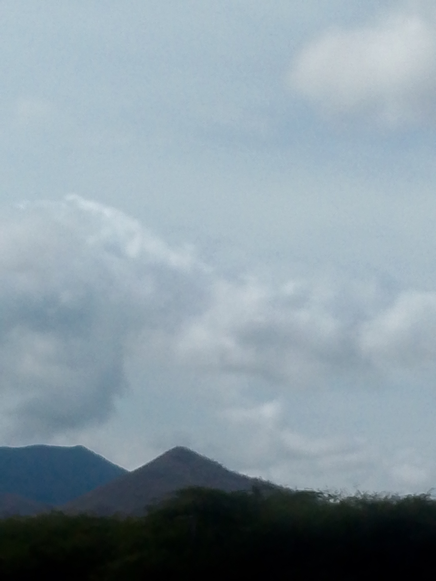 The image shows a landscape with mountains, a dark green line of vegetation, and a cloudy sky. Two mountain peaks are visible, one more prominent than the other. The sky is predominantly cloudy, with some patches of lighter blue visible. The vegetation appears dense. The location is in Juan Griego, Venezuela.