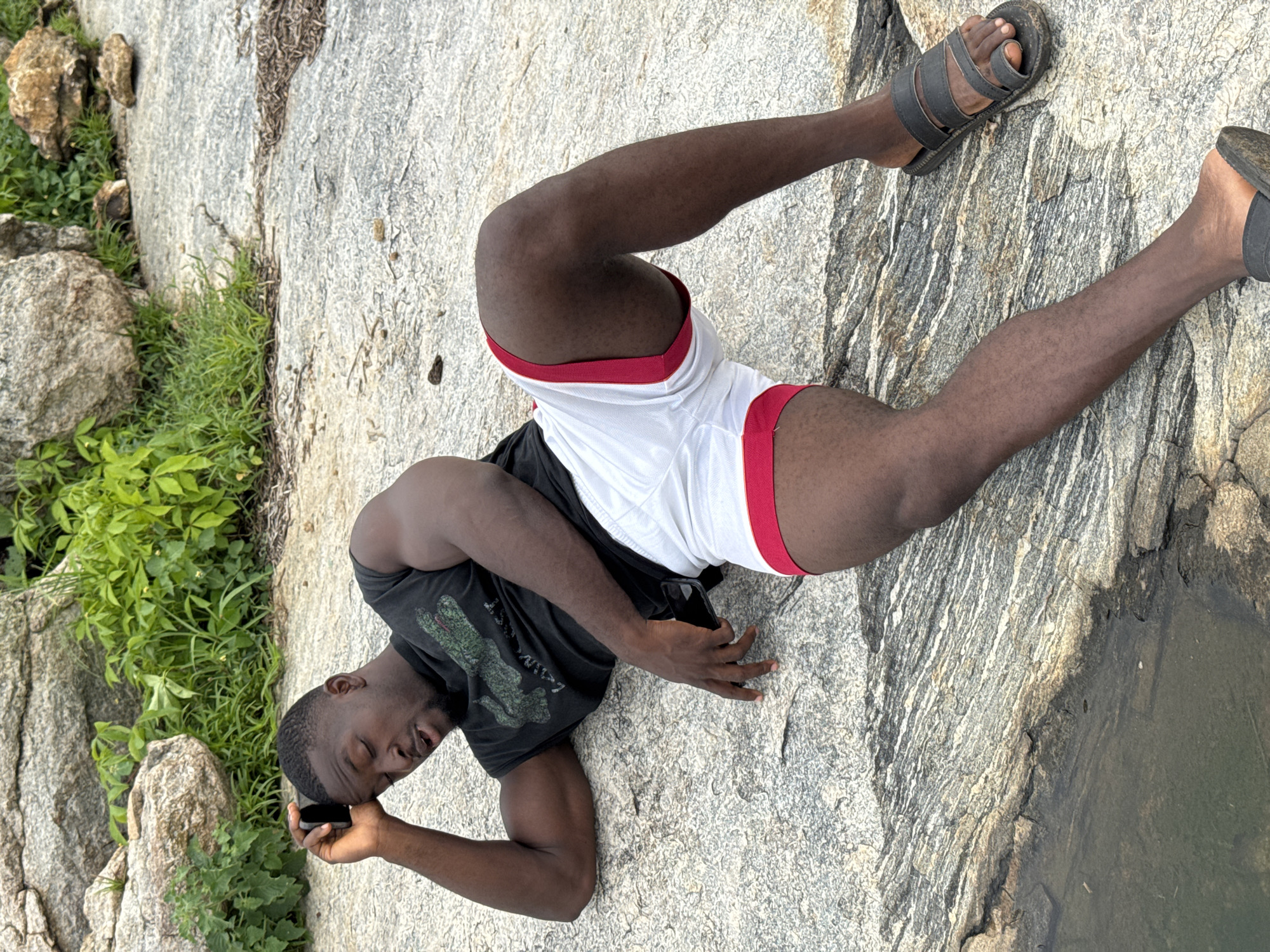 A Black man is lying on a large, grey, textured rock with green foliage and larger rocks in the background. He is wearing a black t-shirt with a green alligator graphic and white shorts with red trim. He is holding a black cell phone to his right ear with his right hand, his eyes are closed, and his mouth is slightly open. His left arm is bent and resting on the rock. He is wearing black sandals on his feet. The rock surface is uneven and appears to be part of a natural outdoor environment, possibly near a body of water as suggested by a dark, wet area to the lower left of the frame. The location is the city of Akwanga, Nigeria.