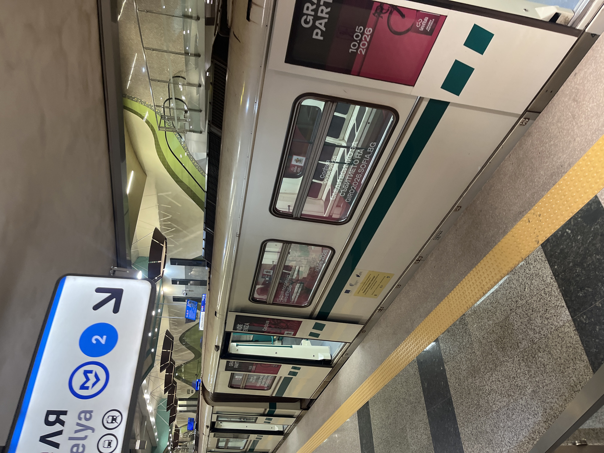 The media file, presented with a rotated orientation, captures a modern underground subway station in Sofia, Bulgaria. A white subway train with a prominent green stripe along its side is stopped at the platform, with one of its doors open, revealing a glimpse of the interior. The platform features a patterned floor of light and dark grey granite tiles, bordered by a yellow tactile safety strip. The station's architecture is characterized by smooth, curving walls, some accented with a light green color, and transparent glass barriers in the background.
Advertisements are prominently displayed on the train's exterior. A large red-and-white ad near the top of the train features a bicycle graphic and the text "GRAND PARTENZA" along with the date "10.05.2026" and "Giro d'Italia". Another ad, visible through a train window and on the side of the train, explicitly states "София посреща Giro d'Italia" (Sofia welcomes Giro d'Italia) and "10.05.2026", with a web address "GIRO2026.SOFIA.BG". These details indicate an upcoming cycling event related to the Giro d'Italia in Sofia in 2026.
On the left side of the frame, a blue-and-white subway sign is partially visible. It displays an arrow pointing upwards, the number '2' within a blue circle, and the stylized 'M' logo for the metro system, indicating Line 2. The Cyrillic text "ЛЯ" and Latin "elya" are also discernible, likely part of the station's name. The scene is illuminated by artificial lighting, typical of an underground subway environment, with no natural light visible.