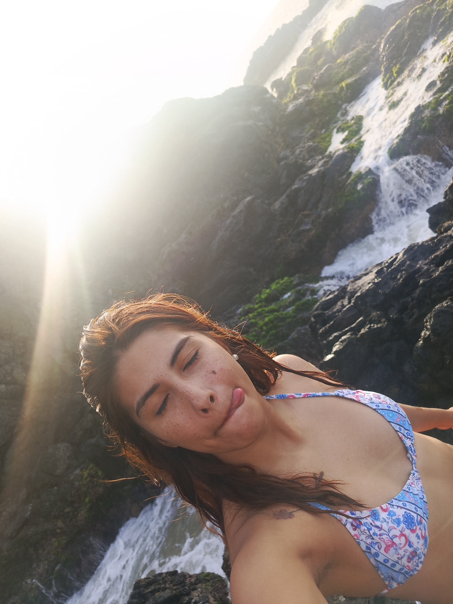 A vibrant outdoor selfie features a young woman amidst a natural, rocky setting. She has closed eyes and her tongue slightly out, conveying a playful expression, possibly squinting from the bright sun. Her hair is brown, and her face shows natural freckles. She is wearing a light blue and white bikini top with a patterned design, possibly floral or paisley.
Behind her, a natural waterfall cascades over dark, rugged rocks. Green moss or algae clings to some of the rocks, adding texture and color to the scene. The water appears white and foamy as it rushes down. The sun is intensely bright in the upper left portion of the image, creating significant lens flare and overexposure in that area, indicating a strong sun source during daytime. This bright light illuminates the woman's hair and part of the background. The scene suggests a wild, untamed environment, likely a coastal or mountainous area in Pedro González, Venezuela, known for its natural beauty. No visible text is present. The overall mood is one of enjoyment and connection with nature.