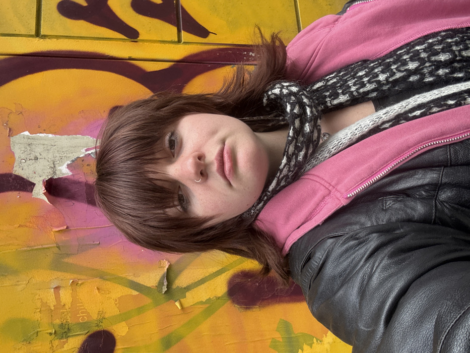 A young woman with brown hair and a nose ring poses against a graffiti-covered wall featuring yellow, pink, and dark brown colors. She wears a black leather jacket over a pink hooded sweatshirt and a patterned scarf. The background is a mix of graffiti art and peeling paint, providing a textured backdrop to the subject's selfie. The camera angle is slightly tilted, focusing on her face and upper body.