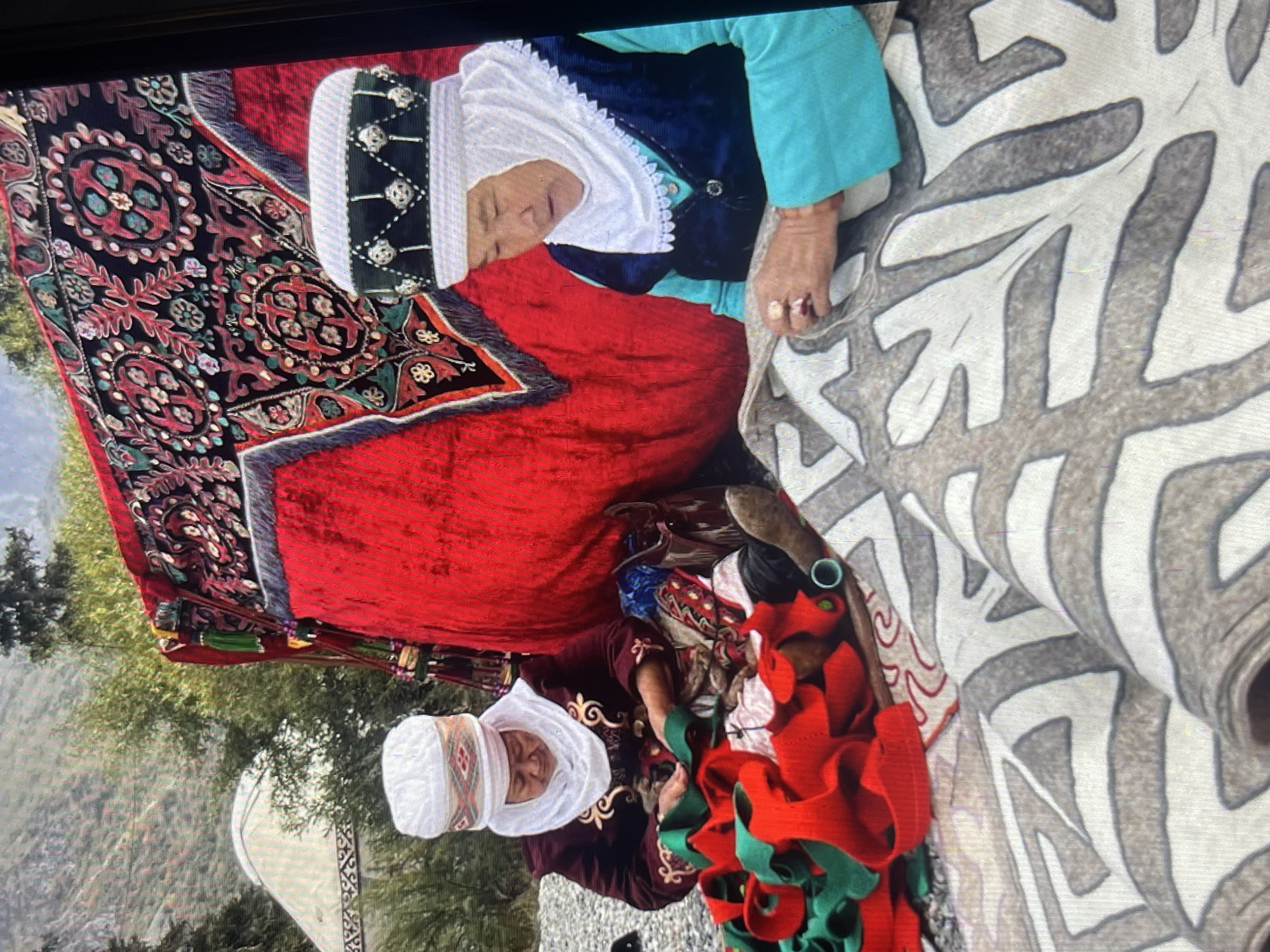 The image shows two Kyrgyz women engaged in textile work in a semi-outdoor setting, possibly near a yurt. The woman on the left, wearing a maroon jacket and a white headscarf, is working with a red and green fabric. The woman on the right, dressed in a blue vest over a turquoise top and a white headscarf, is also working on a textile, which appears to be a rug with a grey and white patterned design. A large, ornately decorated red fabric structure, likely a tent or canopy, is behind them, featuring intricate red, black, and green patterns. In the background, a white yurt is visible, along with lush greenery and hills, suggesting a rural or mountainous environment near Bishkek, Kyrgyzstan. The overall scene depicts traditional Kyrgyz craftsmanship.