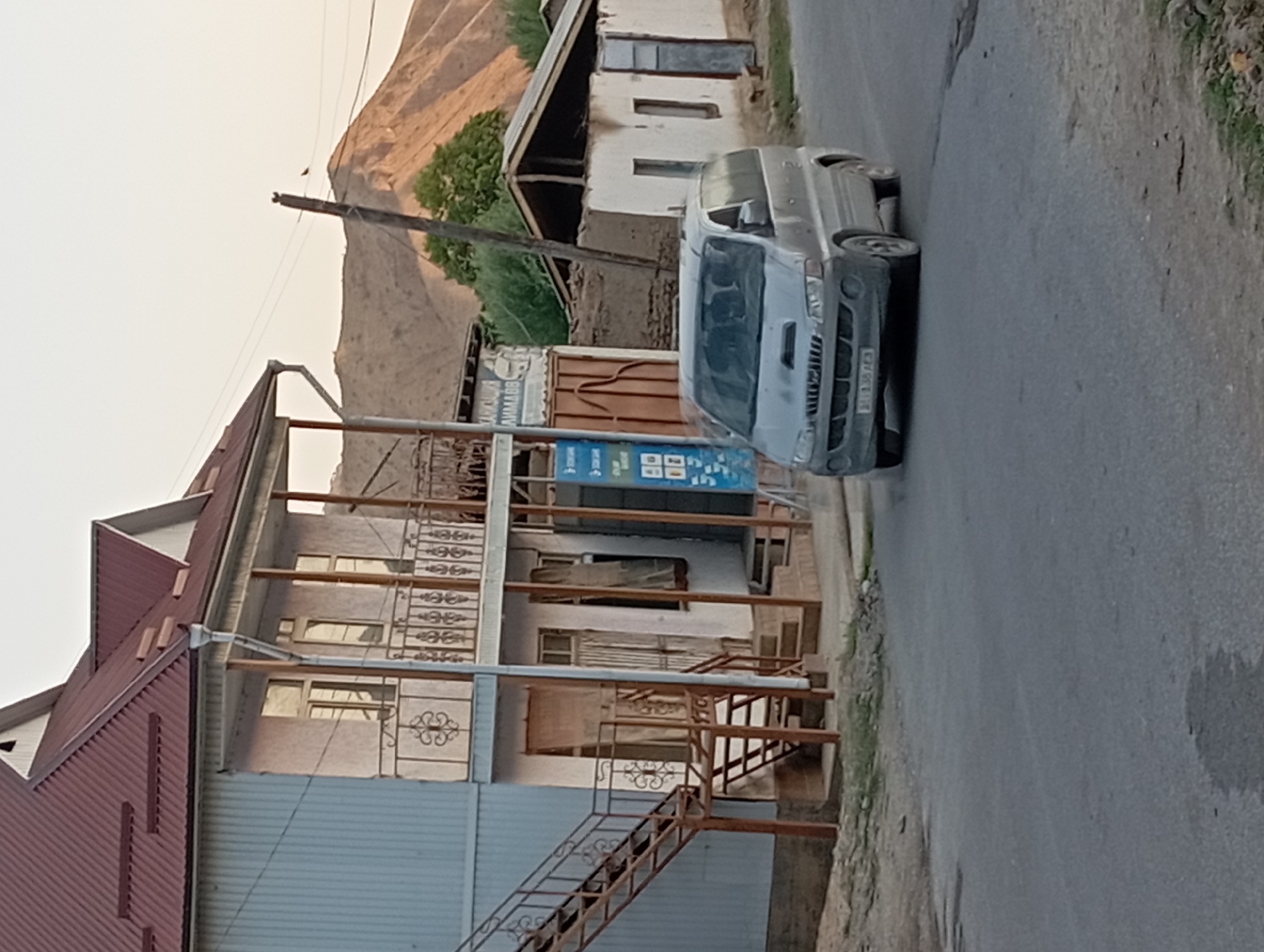 The image captures a street scene in Ak-Suu, Kyrgyzstan, during daylight. In the foreground, a gray minivan, possibly a Hyundai Starex, is parked on an asphalt road. The minivan has its headlights on and a license plate that is partially obscured. To the left, a two-story building with a corrugated metal exterior and a dark red metal roof is visible. This building features balconies with decorative metal railings and an external metal staircase leading to the upper floor. One of the doors on the ground floor is covered by a light-colored cloth. Adjacent to this building, a smaller, older structure with a white facade and a sloped roof is situated. In the background, rolling hills with dry, barren slopes dominate the landscape, with sparse green trees dotting the scene. A utility pole with wires runs across the upper portion of the image, with two small birds perched on the wires near the top of the pole. The sky is a pale, hazy orange, suggesting it might be late afternoon or early morning.
