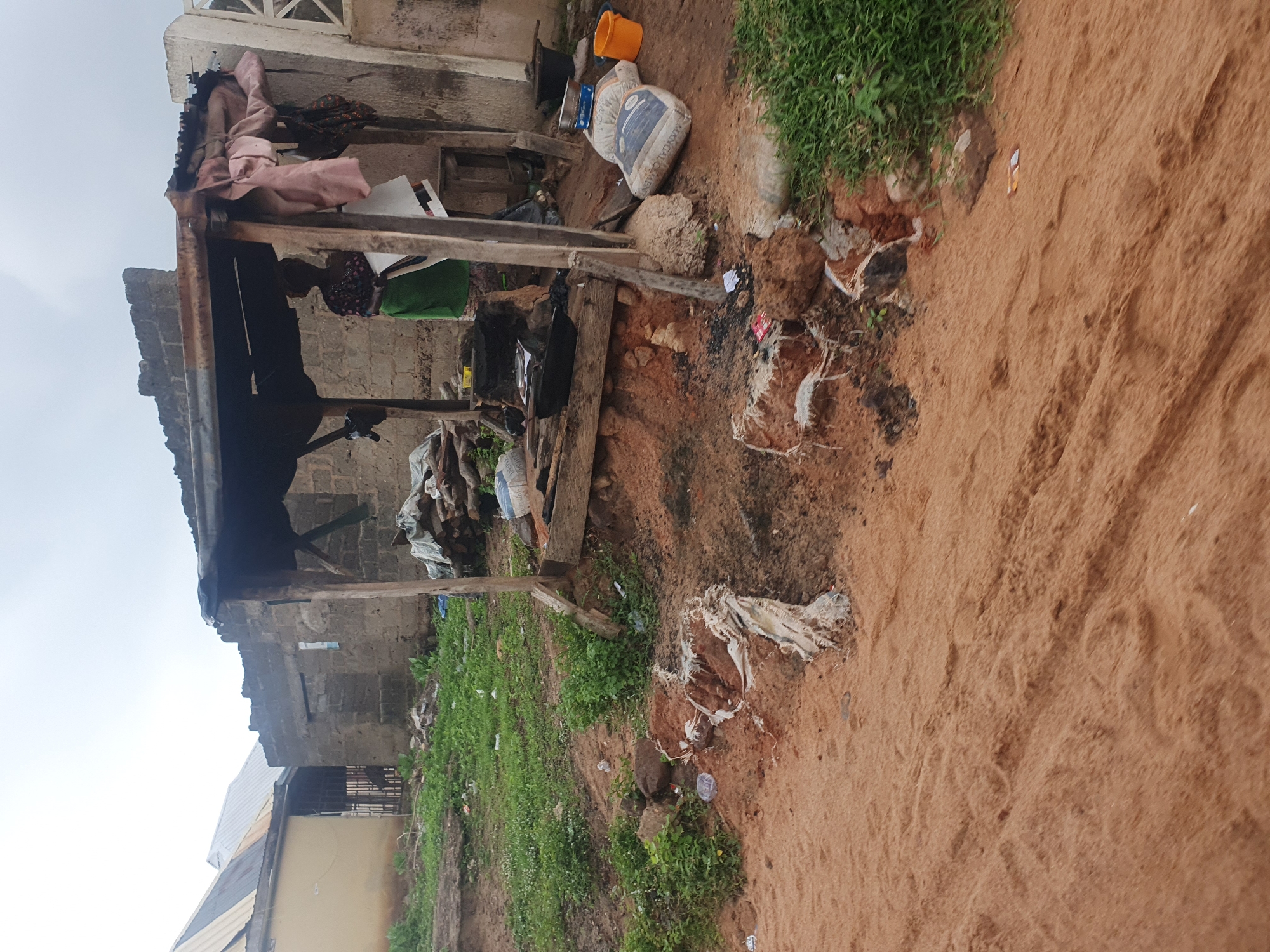 The image depicts an outdoor scene at what appears to be a market stall or roadside vendor area in Lokoja, Nigeria. The foreground is dominated by reddish-brown sand with tire tracks and some scattered debris. In the midground, there is a roughly constructed shelter with a corrugated metal roof, supported by wooden posts. Inside and around the shelter, there are various items. A woman, dressed in a patterned top and a green skirt, is partially visible within the shelter. She is holding what appears to be a large white sheet or piece of fabric.
Objects in the scene include sacks, possibly of cement or similar material, stacked near the shelter. One sack has visible text reading "MAXI-CAL CIMENT". There are also several black cooking pots or basins, a silver sieve, and a bright orange bucket. Piles of wood or debris are scattered around the area. The ground is uneven, with patches of grass and exposed earth, suggesting a less developed or possibly rural part of the city. In the background, there are buildings constructed from cinder blocks, some with corrugated metal roofs. The sky is overcast, indicating cloudy weather. The overall impression is one of a functional, albeit somewhat unkempt, commercial space in a Nigerian urban environment.
