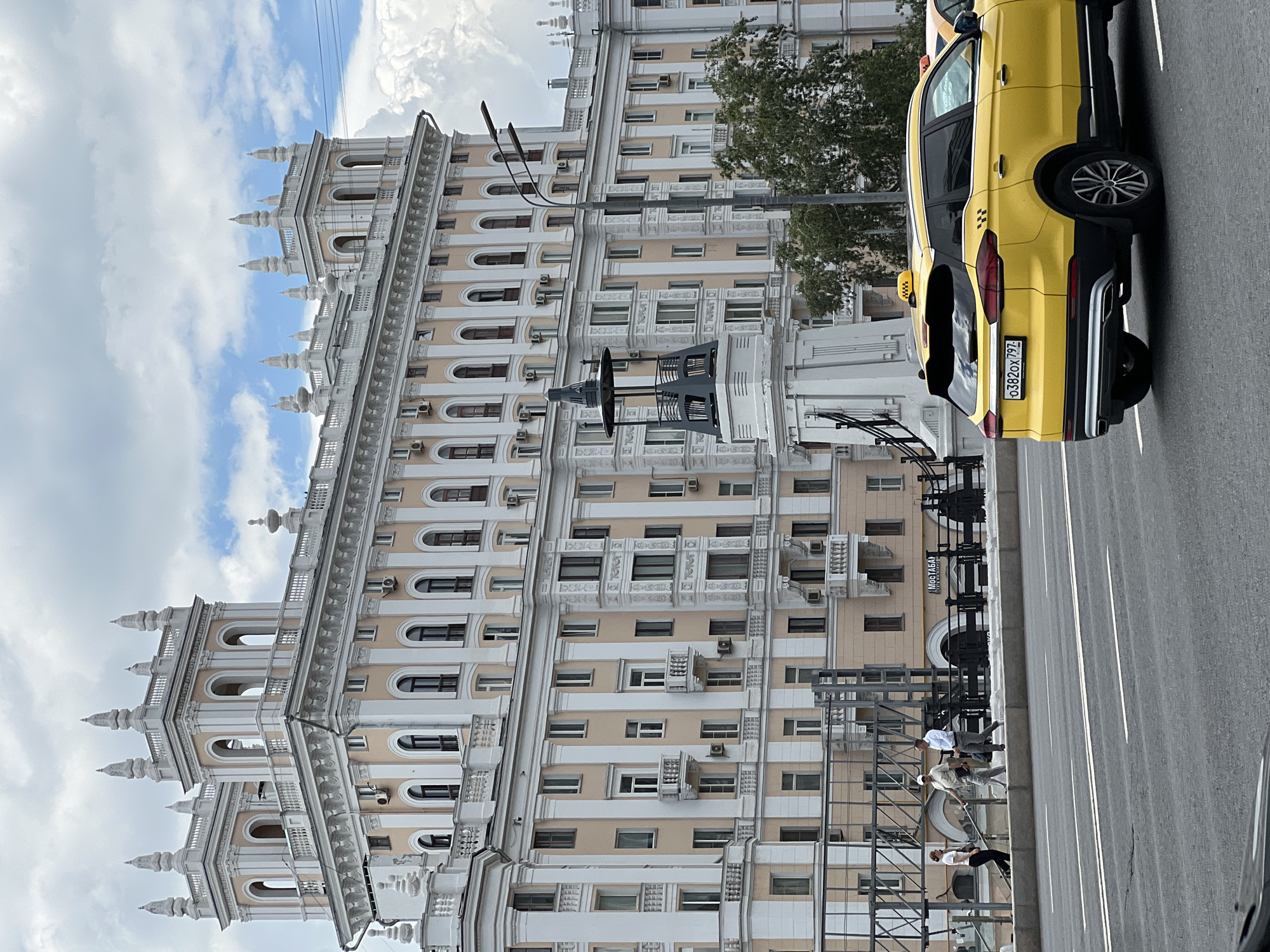 The image shows a street scene in Moscow, Russia, with a large, ornate building in the background. The building features multiple stories, white columns, arched windows, and decorative spires on its roof. In the foreground, a yellow taxi with a checkerboard pattern and Russian license plates is visible on the road. Three pedestrians are walking on a sidewalk in front of the building, which is separated from the road by a concrete barrier. The sky is partly cloudy. The building appears to be a Stalinist Empire style skyscraper.