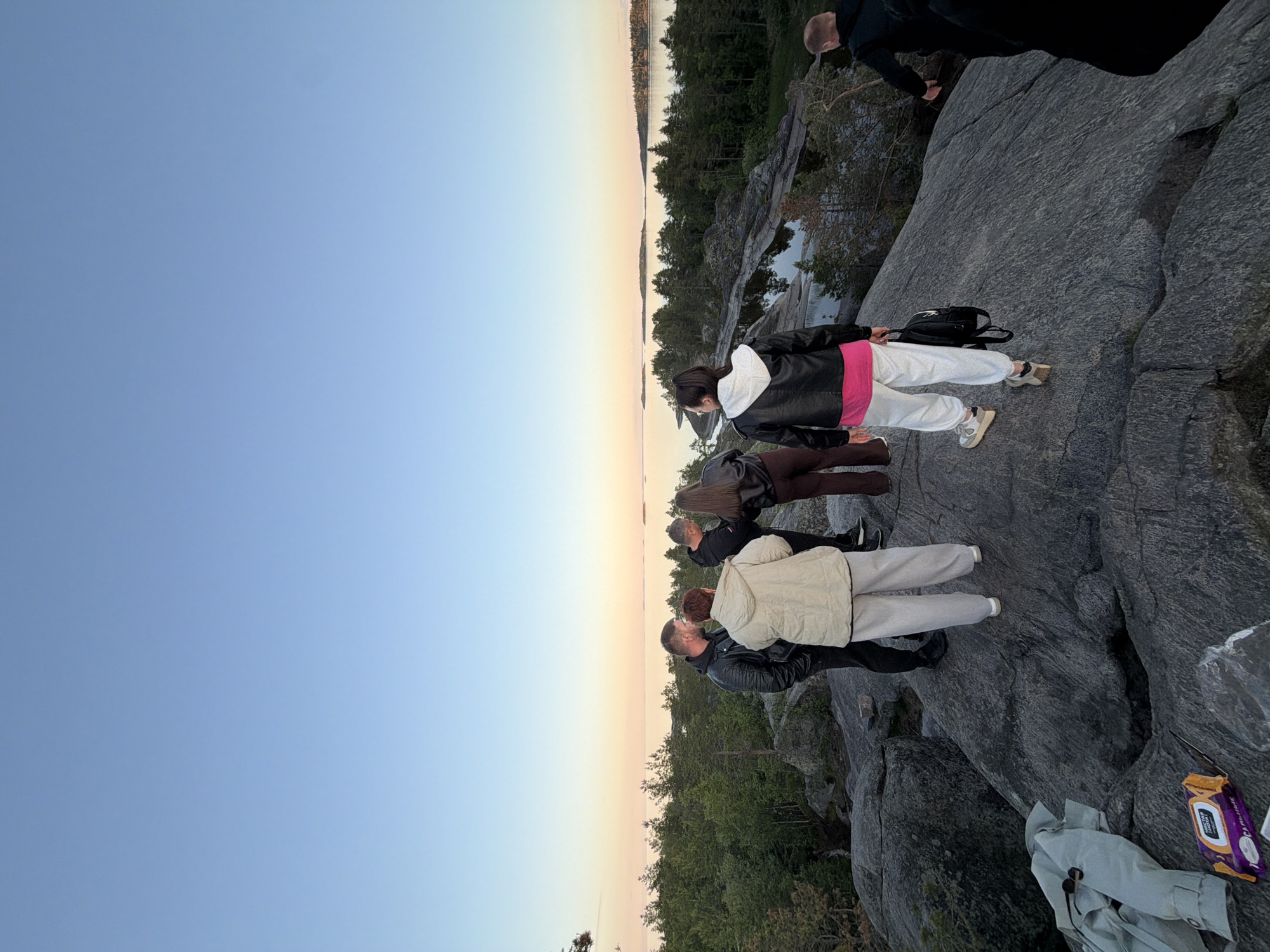 A group of six individuals are gathered on a rocky outcrop overlooking a serene body of water bordered by a dense forest. The sky displays a gradient of soft blues and pinks, indicating either sunrise or sunset. The people, dressed in casual attire, appear to be engaged in conversation or observing the scenery. Some are facing the water, while others are turned towards each other.
One individual in the foreground, facing away from the camera, wears white pants and a black jacket with a pink top underneath. They are holding a black backpack. To their left, a person in a light-colored jacket and white pants stands facing them. Two other individuals, a man in a black leather jacket and a woman in a black jacket, are in conversation to the left of the group. Further to the right, a man in a black hoodie is walking away from the camera, and another person, also in dark clothing, is partially visible on the far right.
On the rock in the foreground, a light-colored jacket and a package of wet wipes are visible. The terrain is rugged and uneven, typical of a natural landscape with large boulders and sparse vegetation. The water appears calm, reflecting the soft hues of the sky. The forest is a mix of evergreen and deciduous trees, creating a lush green backdrop against the water. The overall scene conveys a sense of tranquility and companionship in a natural setting.