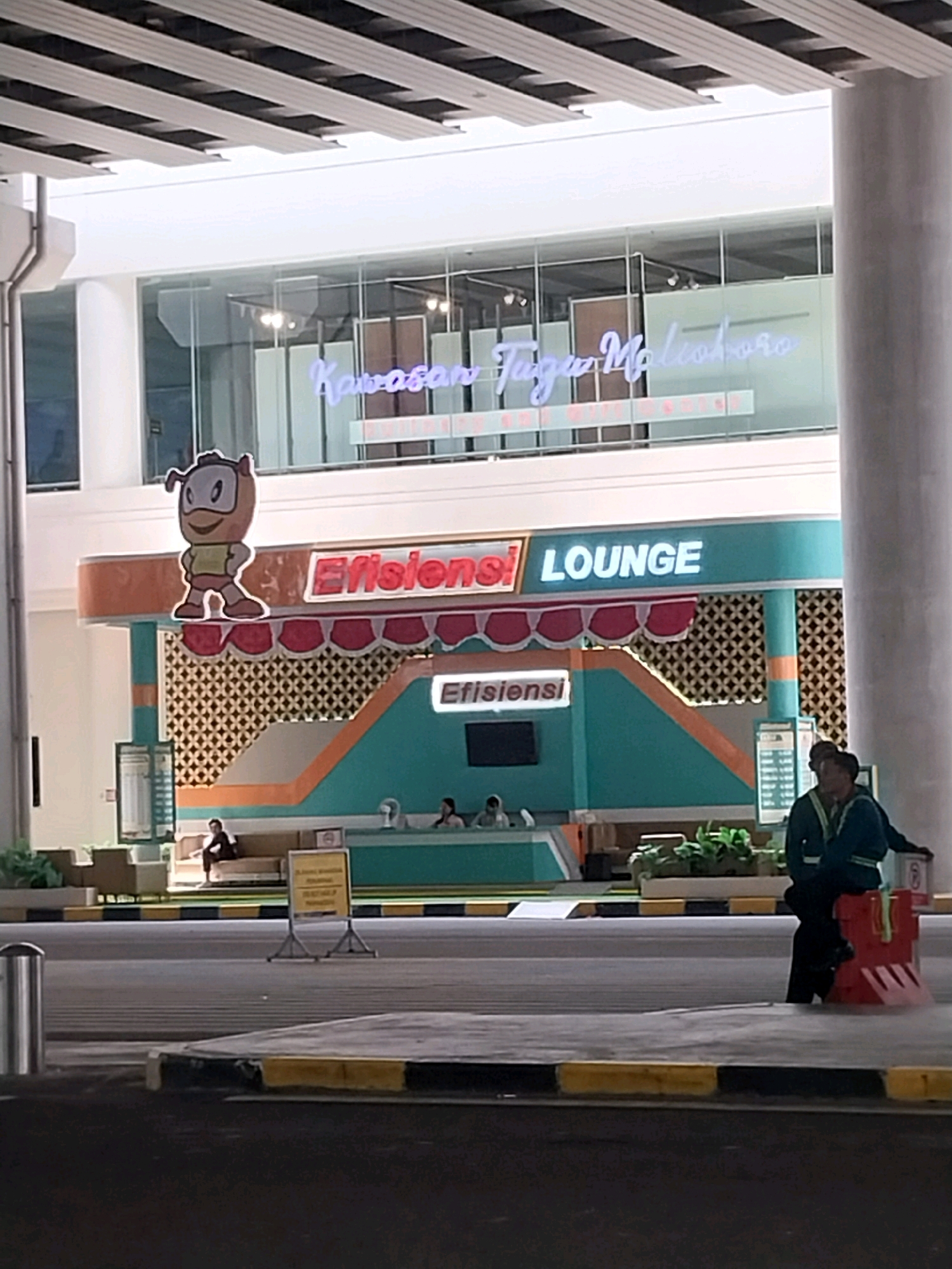 In Palihan, Indonesia, an outdoor lounge area is visible. Above the lounge, a neon sign in Indonesian reads "Kawasan Tugu Malioboro". The lounge itself is decorated with a teal and orange color scheme, featuring a cutout mascot character and the words "Efisiensi LOUNGE" in illuminated lettering. Inside, several people are seated. Two individuals, presumably security personnel, are standing outside the lounge. One is wearing a high-visibility jacket over a dark uniform, and the other is partially visible behind them. The area is demarcated by yellow and black striped barriers.