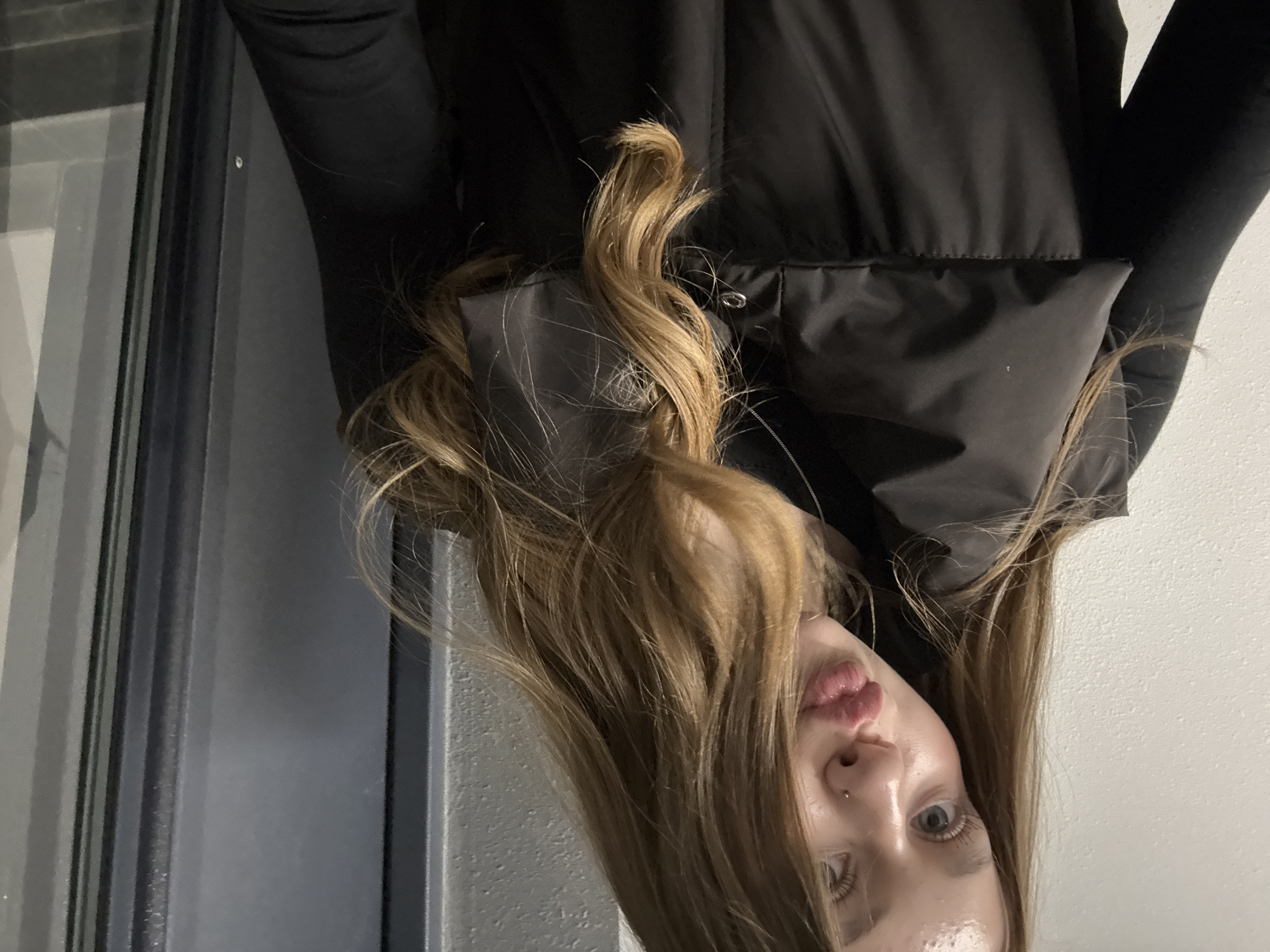 A woman with long, light brown hair is posing for a picture. She is wearing a black puffer jacket. The background includes a glass window and a white wall. The image is taken at an angle, with the woman's head tilted. No location information is discernable.
