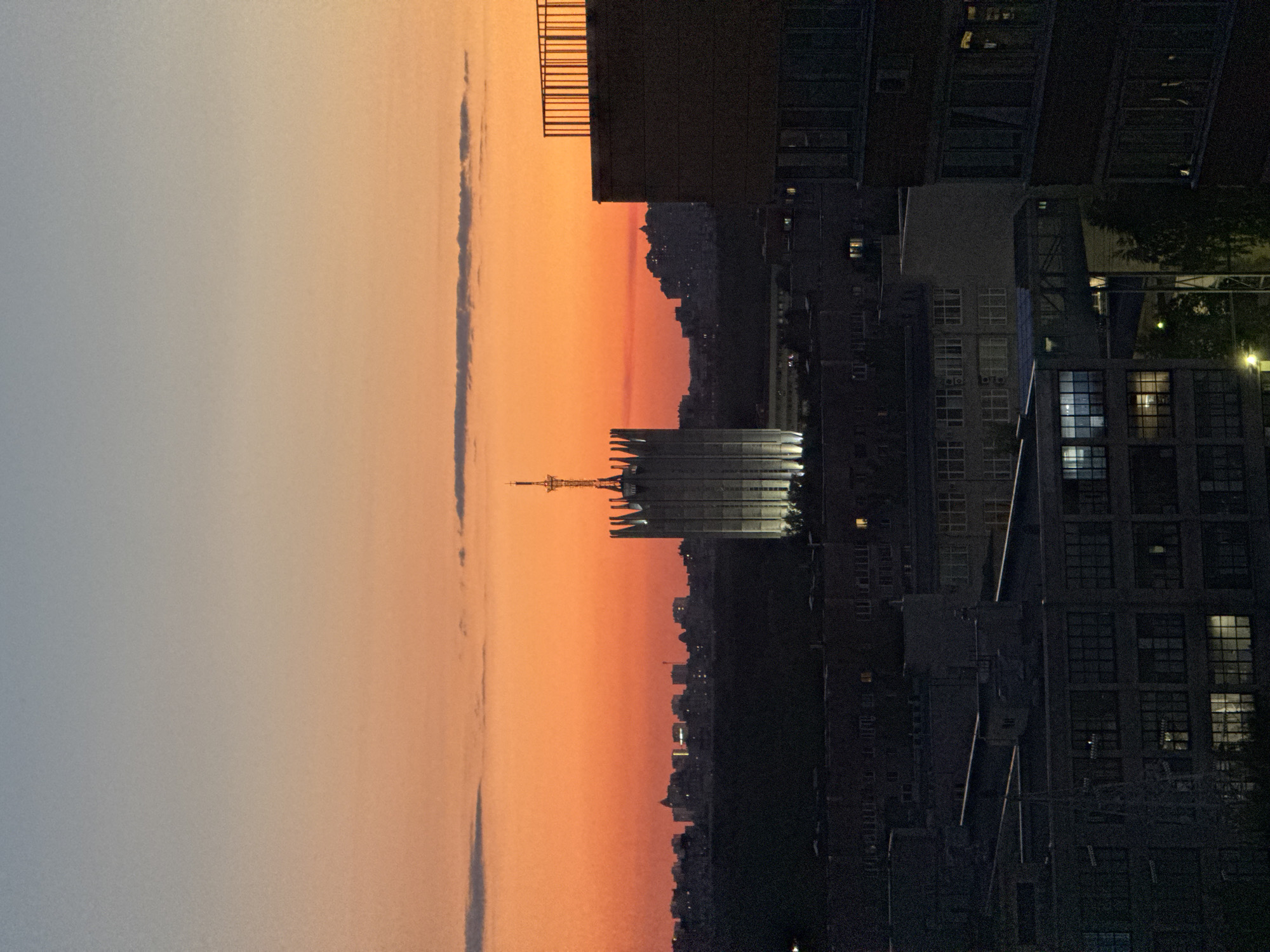 An urban landscape in Unknown, Russia, is depicted at dusk or sunset. The sky transitions from a pale blue at the top to a vibrant orange-red at the horizon, featuring several narrow, dark cloud formations. A prominent, tall building occupies the central midground, characterized by a complex, multi-finned apex and a central spire. This building is illuminated from its base, causing it to stand out against the darker foreground and the bright sky. In the distant background, a silhouette of a city or settlement is visible against the horizon, with scattered points of artificial light. The foreground and midground are dominated by numerous dark, multi-story buildings, consistent with commercial or industrial architecture. Multiple windows within these foreground structures exhibit internal illumination, and isolated ground-level light sources are present. A building on the right side of the frame features a rooftop railing. No individuals or specific actions are discernible within the scene.
