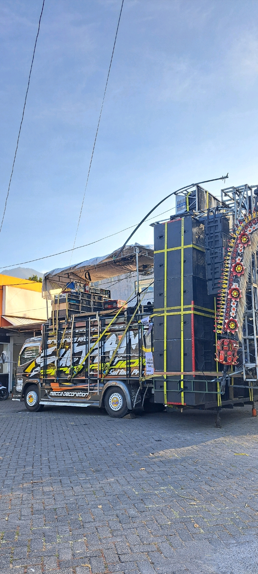 In Prigen, Indonesia, a large truck is parked on a paved area, likely a street or parking lot. The truck is heavily modified and appears to be a sound system vehicle, often used for events or festivals. It is laden with numerous large speakers stacked high on its bed and a metal scaffolding structure extending upwards. The truck itself is adorned with vibrant decals featuring graphics and text, including "GULTAR" and "MECCA DECORATION". Yellow straps are used to secure the speakers to the truck and scaffolding. To the left of the truck, a building with a yellow facade is visible. The sky is a clear, pale blue. The overall scene suggests preparation for or aftermath of an outdoor event.